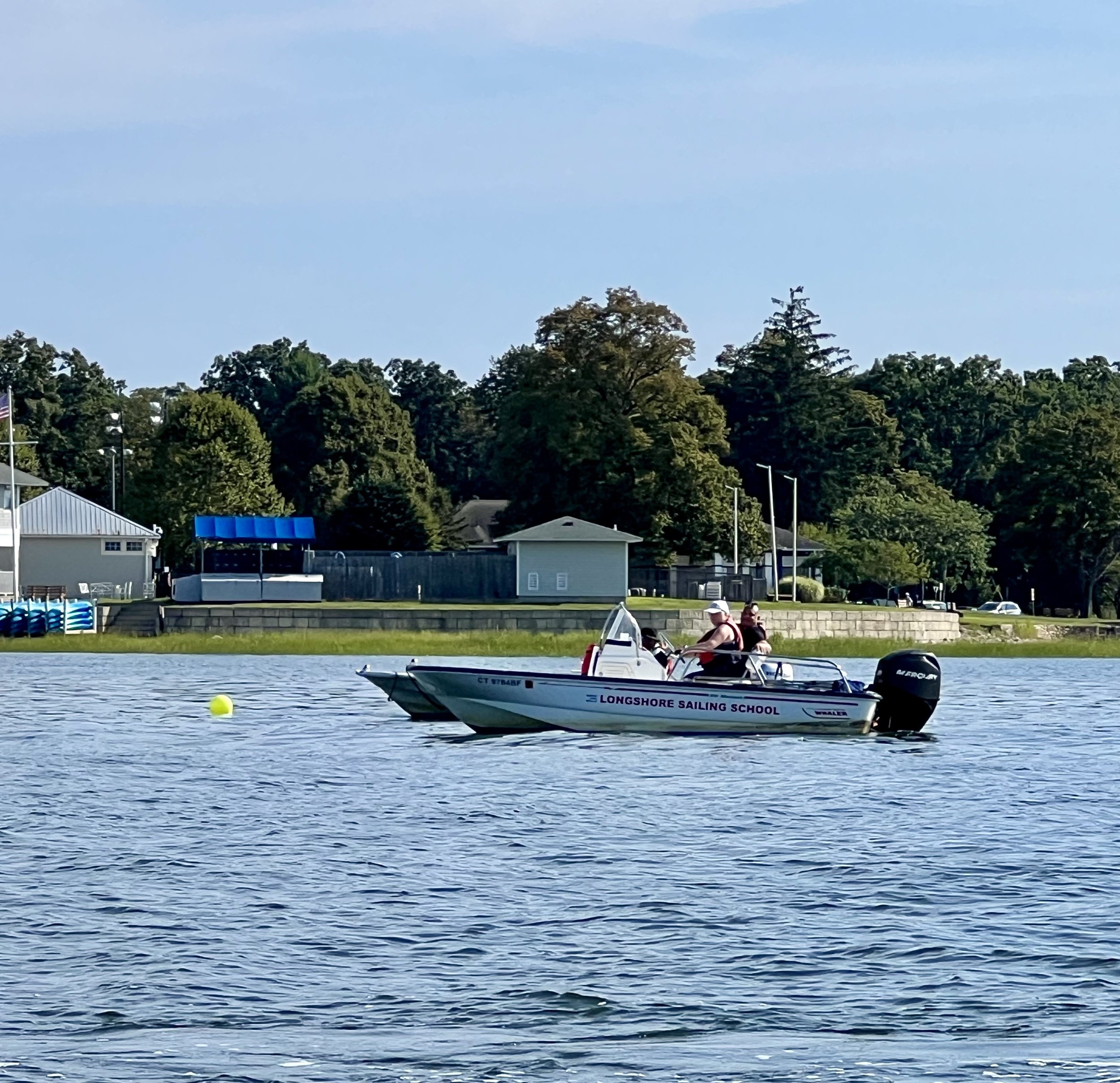 A motorboat from Longshore Sailing School on water near the shore with people on board. Trees and buildings are visible in the background.
