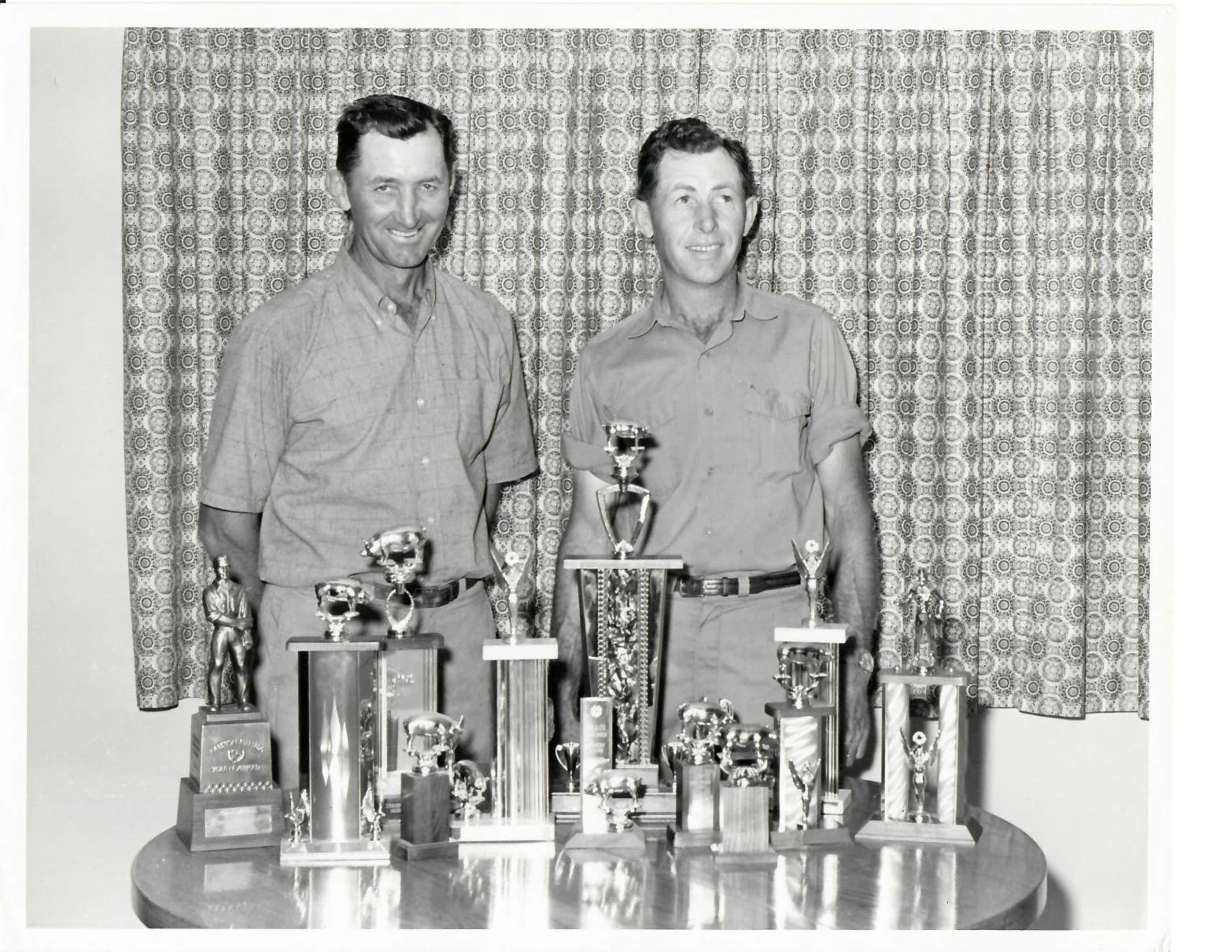 Black and white photo of two men standing behind a table filled with numerous trophies and awards, with patterned curtains in the background.