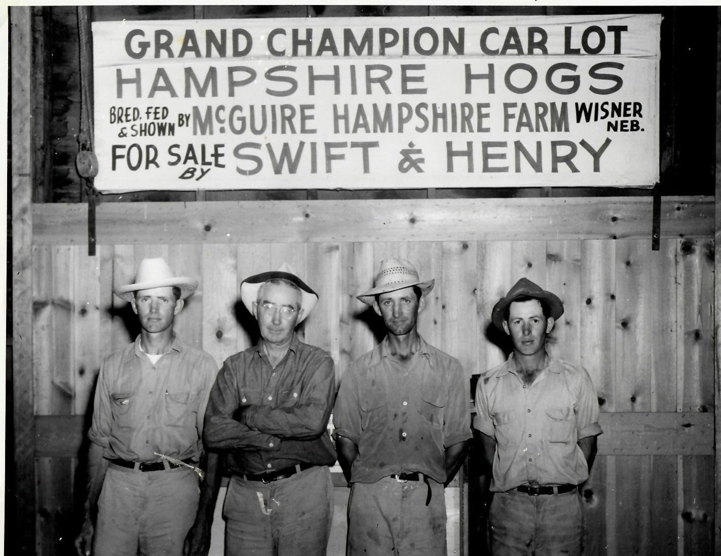 Four men standing in front of a wooden fence and a sign that reads, 'Grand Champion Car Lot Hampshire Hogs bred fed & shown by McGuire Hampshire Farm Wisner Neb. For sale by Swift & Henry.' The men are wearing hats and casual clothing.