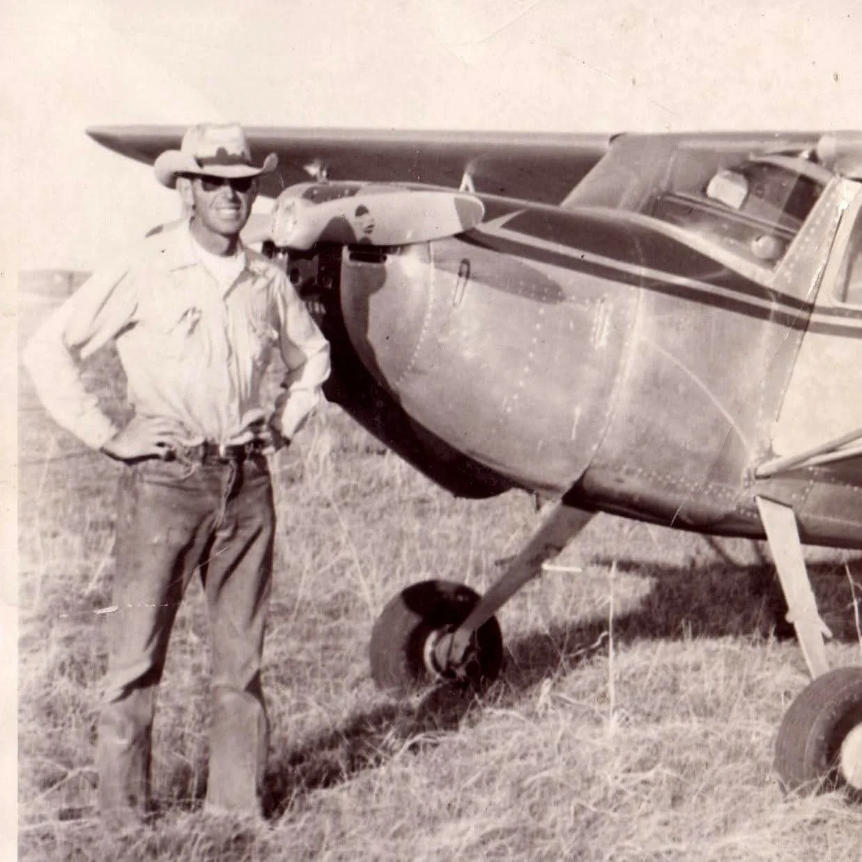 Black and white photo of man standing next to small airplane on grass field, wearing sunglasses, cowboy hat, and casual military-style clothes.