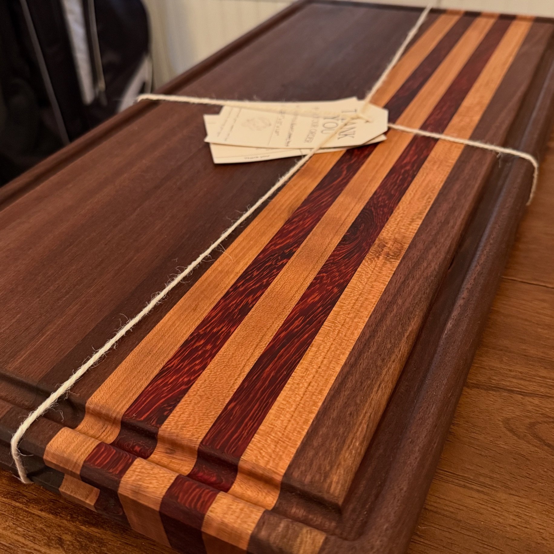 A wooden cutting board with a striped pattern of lighter and darker wood inlays, tied with a piece of string and a paper tag on top.