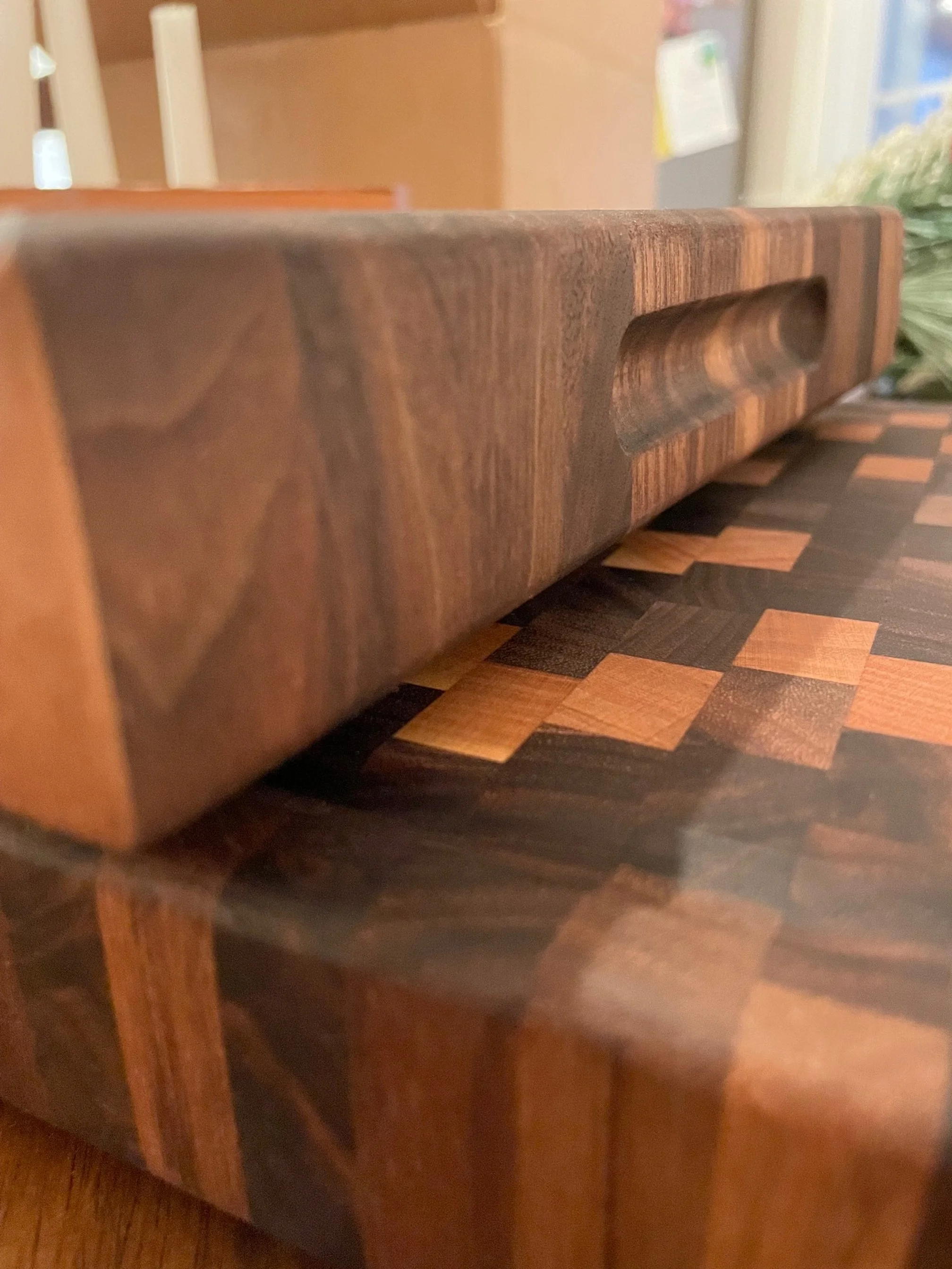 Close-up of a wooden cutting board with a groove for gripping, placed on a butcher block countertop with a checkered pattern of dark and light wood.