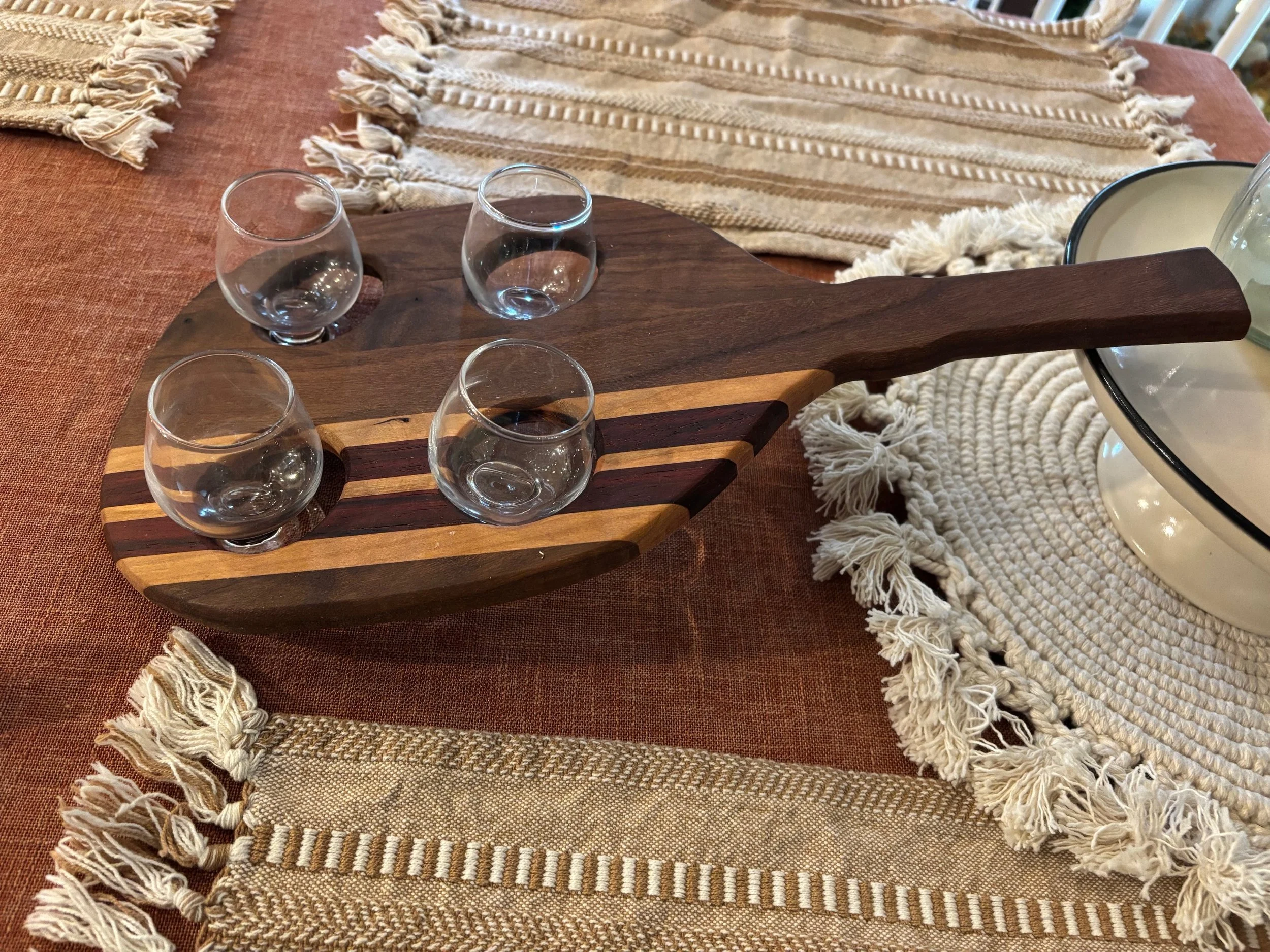 A wooden serving paddle with four empty shot glasses on it, placed on a table with woven napkins and a round woven placemat.