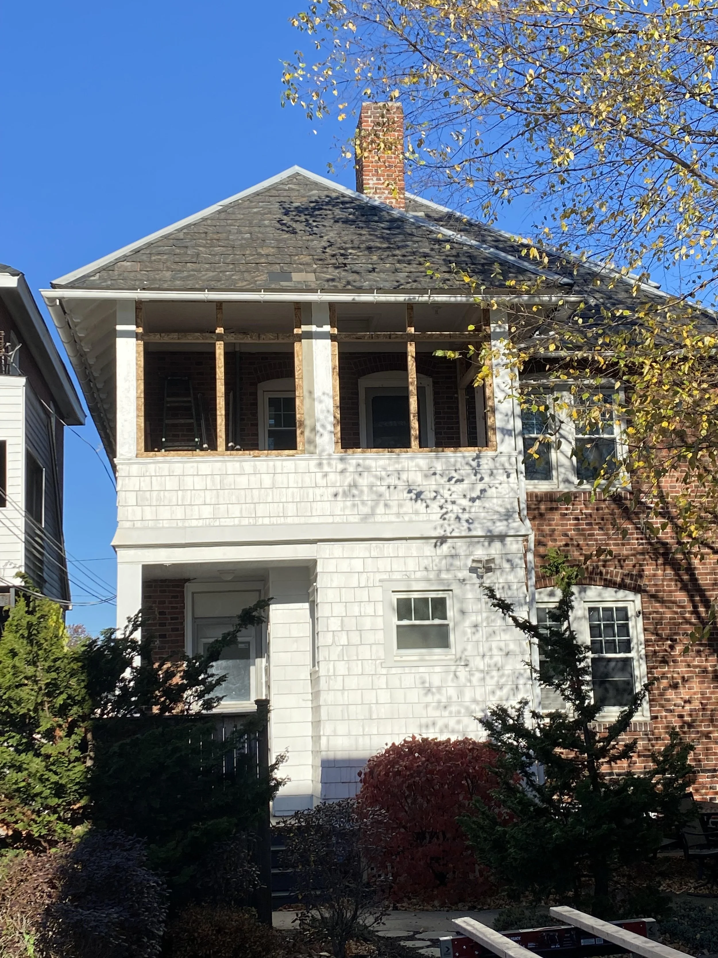 A two-story house under renovation with exposed wooden framing on the upper porch, partially covered with white shingles on the lower level, brick on the right side, and surrounded by trees and bushes, under a clear blue sky.