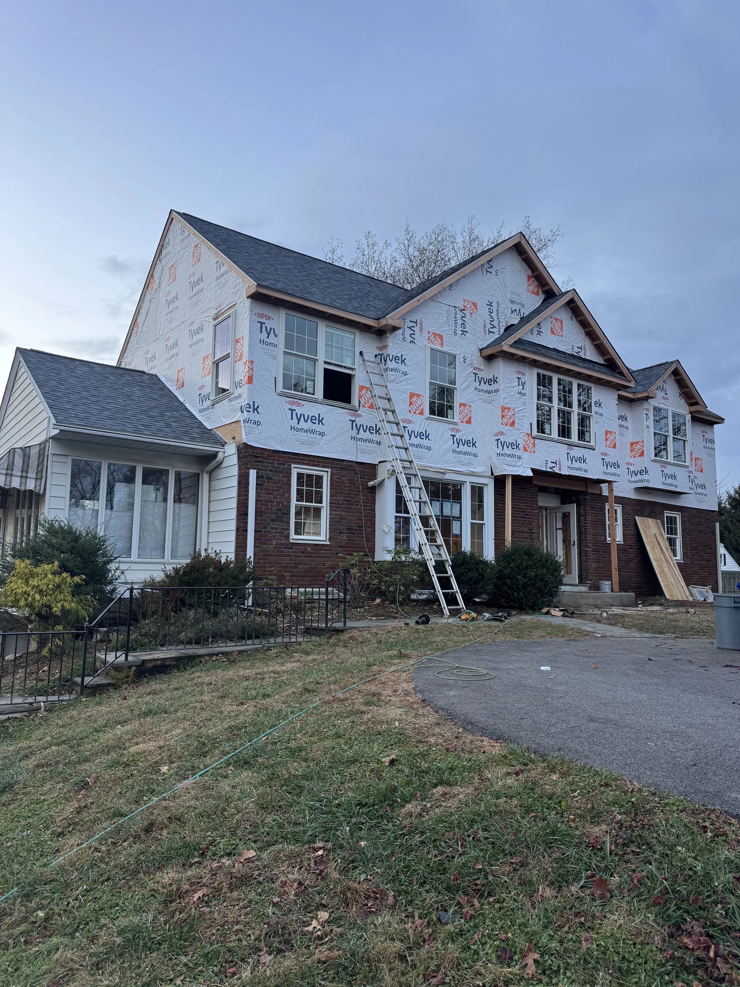 A two-story house under construction with Tyvek HomeWrap on the exterior, some windows installed, and construction equipment and materials around the entrance area.