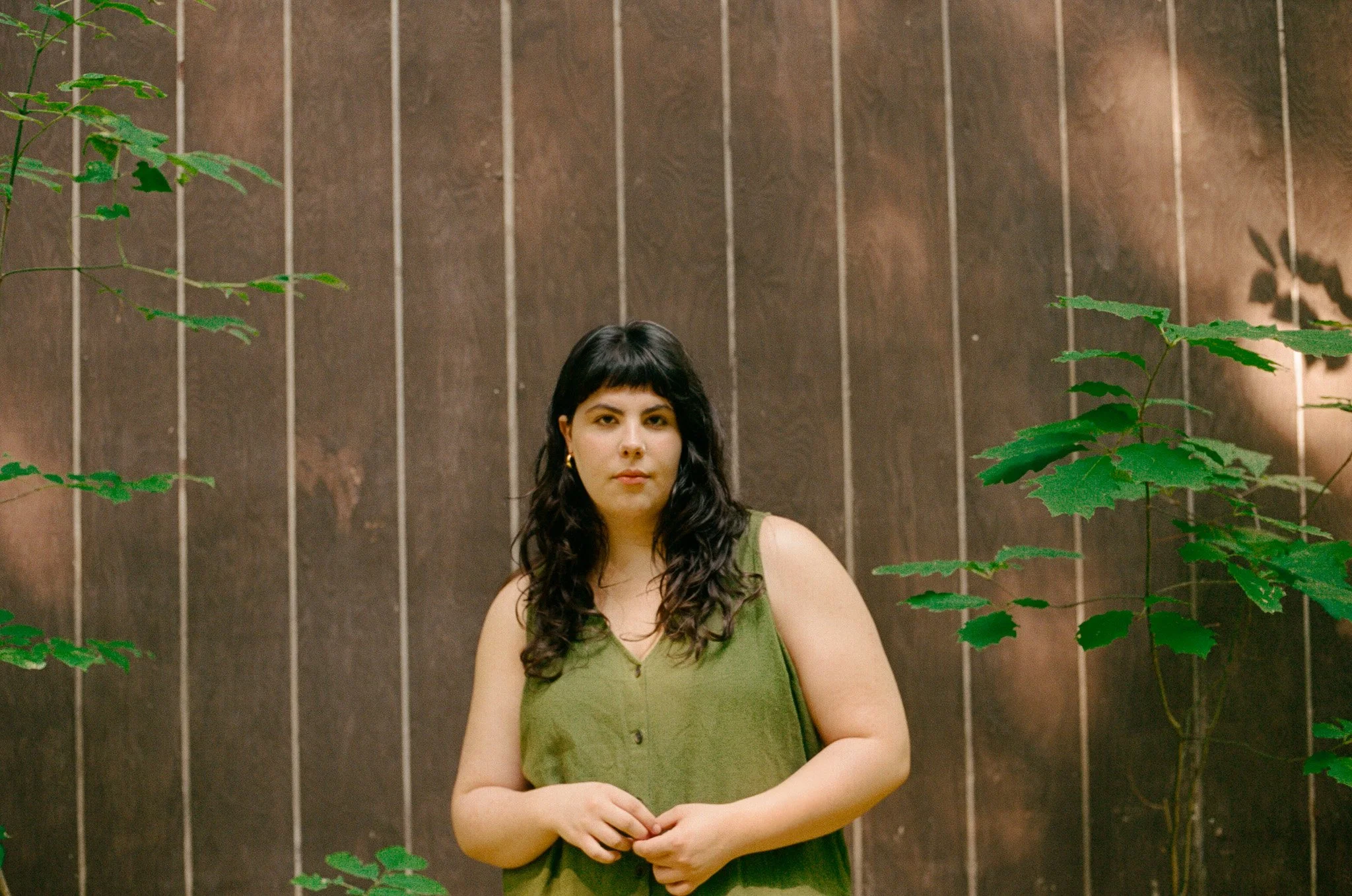 A woman with dark wavy hair and light skin, wearing a green sleeveless top, stands in front of a brown tiled wall, partially framed by green plant leaves.