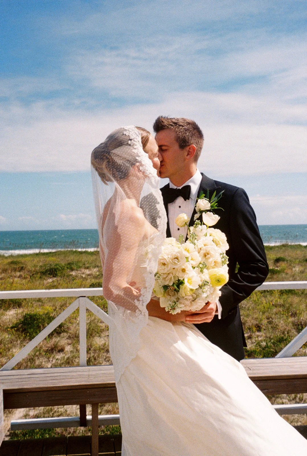A bride and groom sharing a kiss on a beachside wedding photo, with the bride holding a bouquet of white flowers and wearing a veil, while the groom is dressed in a black tuxedo, against a background of blue sky, green grass, and the ocean.