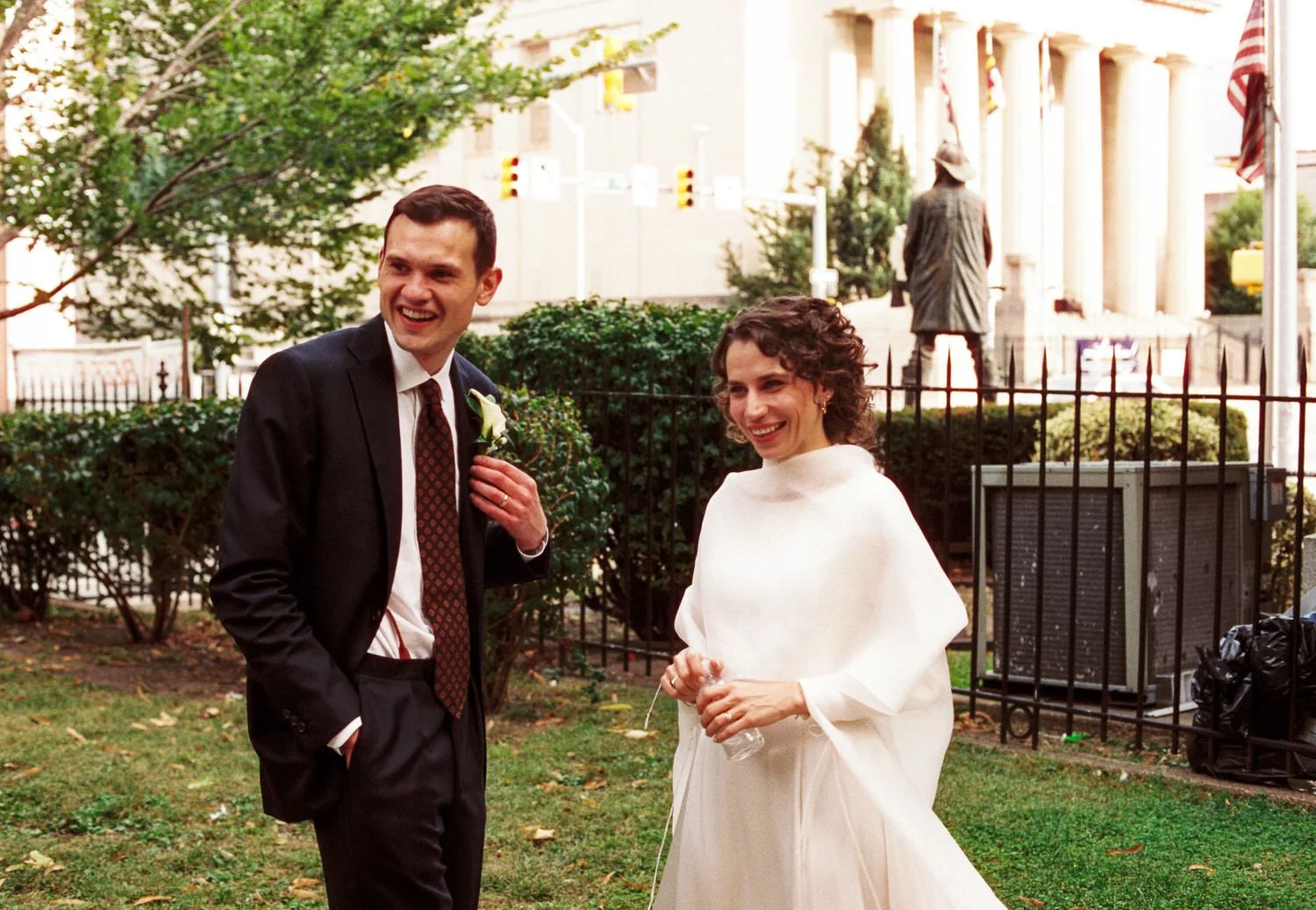 A happy couple, a man in a black suit with a white shirt, red tie, and boutonniere, and a woman in a white dress, are standing outside in a garden, smiling, with a statue and building with columns in the background.