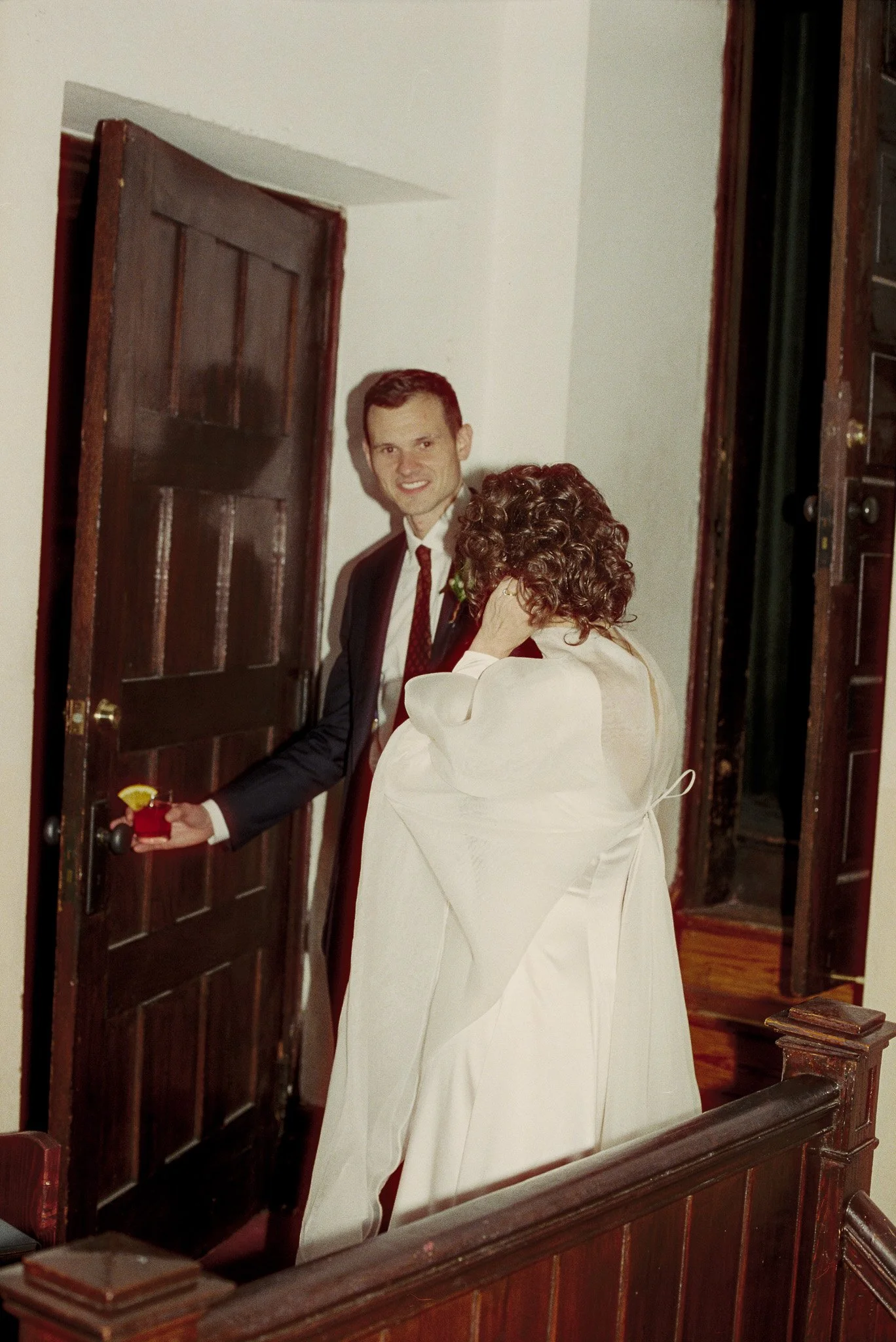 A black-and-white photo of a happy couple, dressed in wedding attire, holding hands and smiling at the camera inside a wooden building with arched ceilings.
