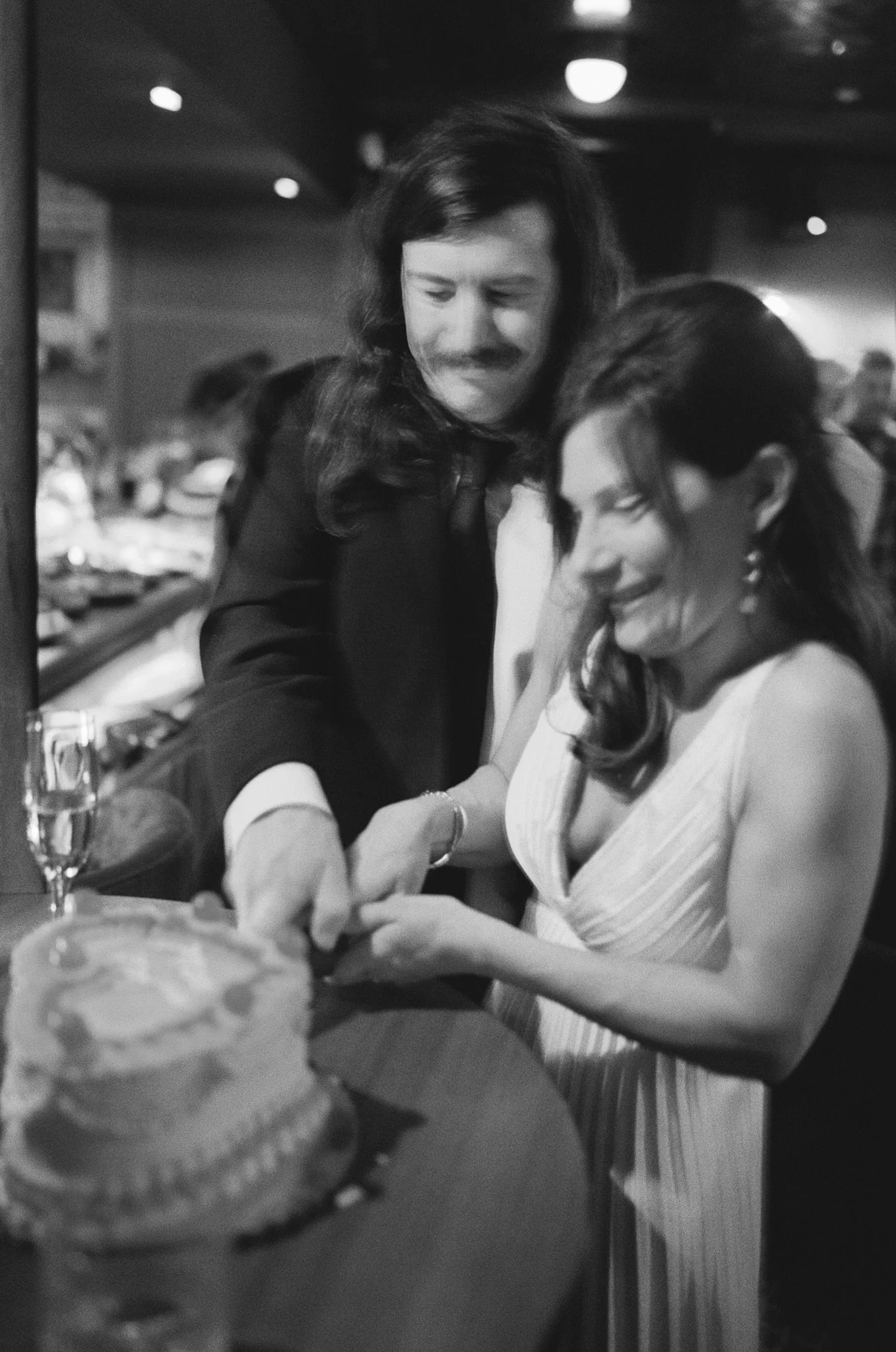 A man and a woman celebrating a special occasion at a restaurant or bar, with a cake and champagne in the foreground, as they smile and talk while looking at a phone.