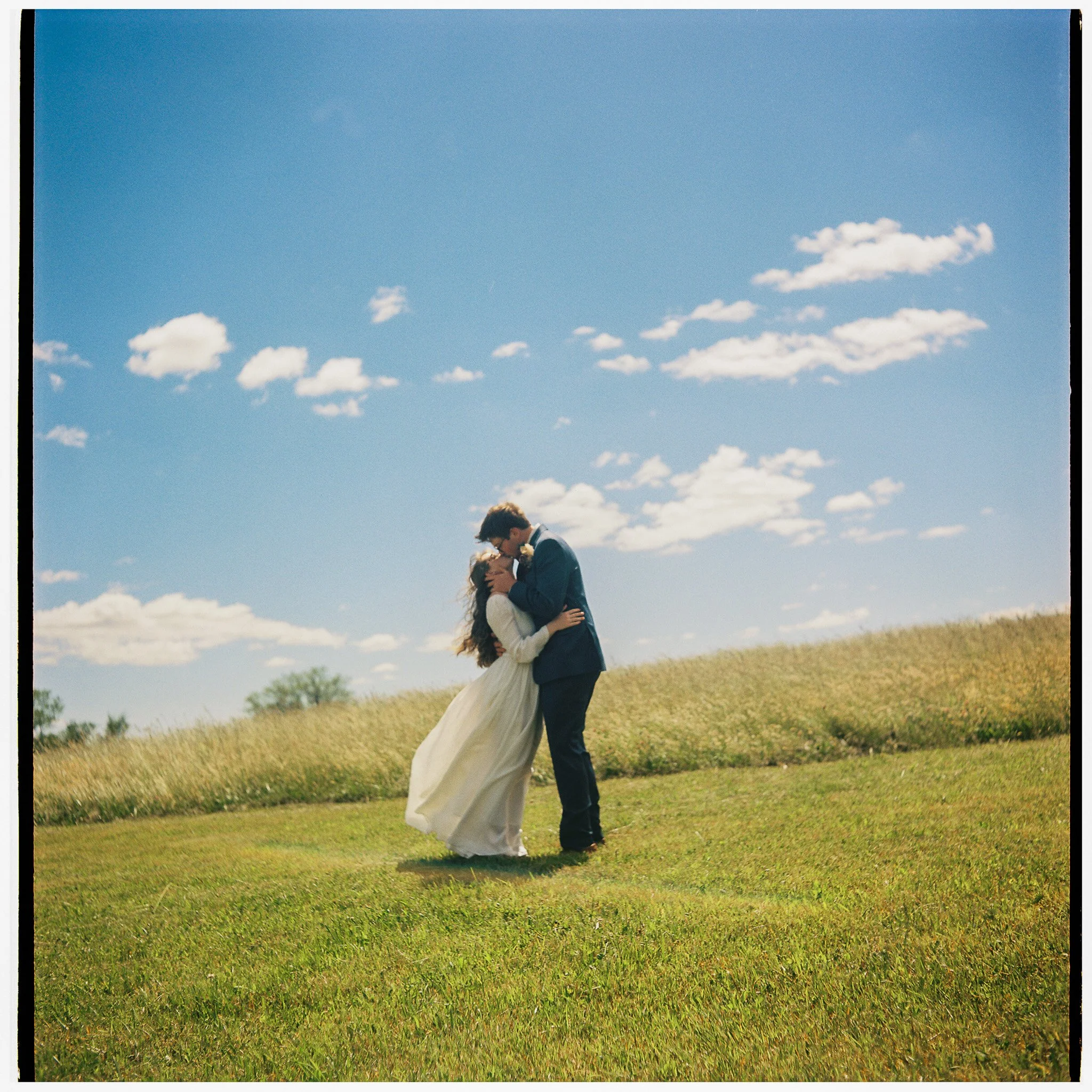 A couple dressed in wedding attire embracing and kissing outdoors in a grassy field under a blue sky with scattered clouds.