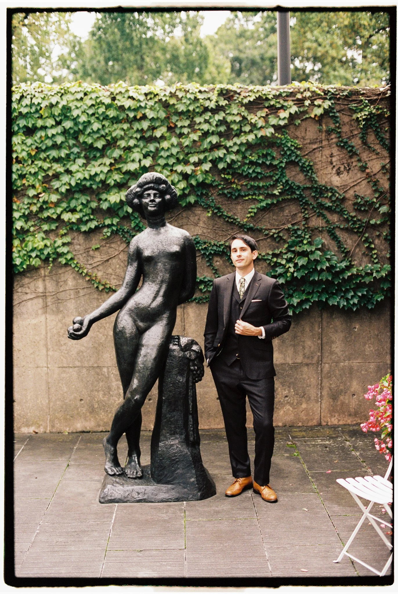 A man in a dark suit standing next to a bronze statue of a woman and a child, outdoors with a green ivy-covered wall in the background.