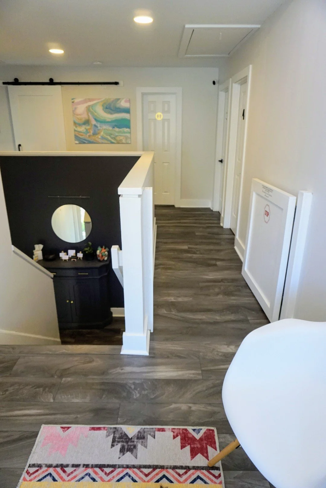Interior view of a hallway featuring hardwood flooring, a staircase with black and white decor, a white round chair, and a colorful abstract painting on the wall.