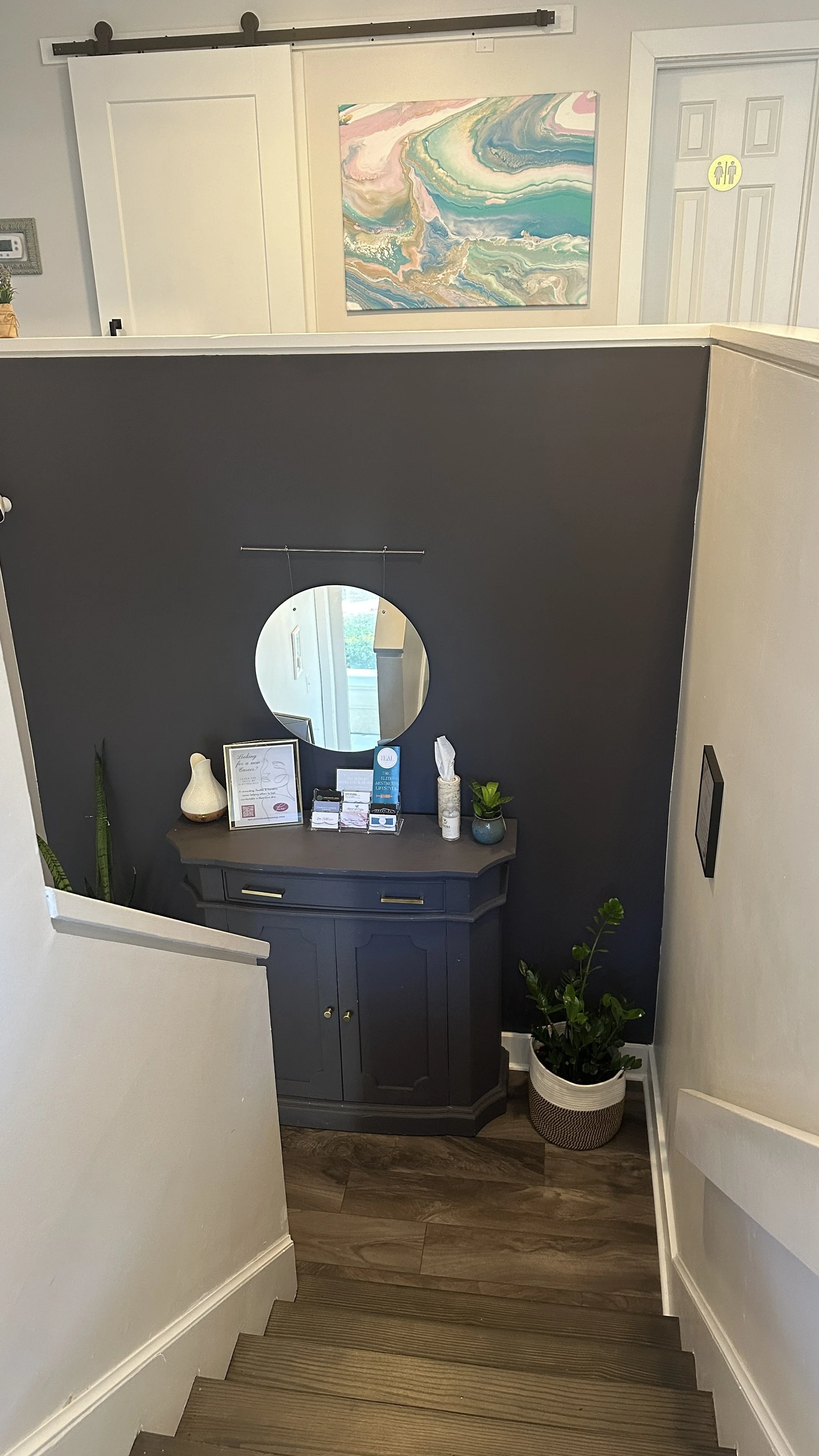 Indoor stairway leading to a small reception area with a dark-colored cabinet, a circular mirror, potted plants, and various items on the cabinet. An abstract colorful painting is on the upper wall, and a door with a restroom sign is visible.