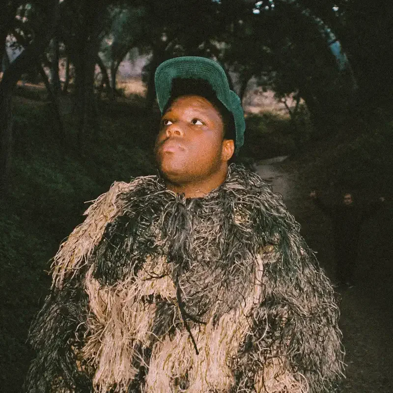 A person wearing a green cap and a large, textured, multicolored fur coat outdoors at dusk or dawn.