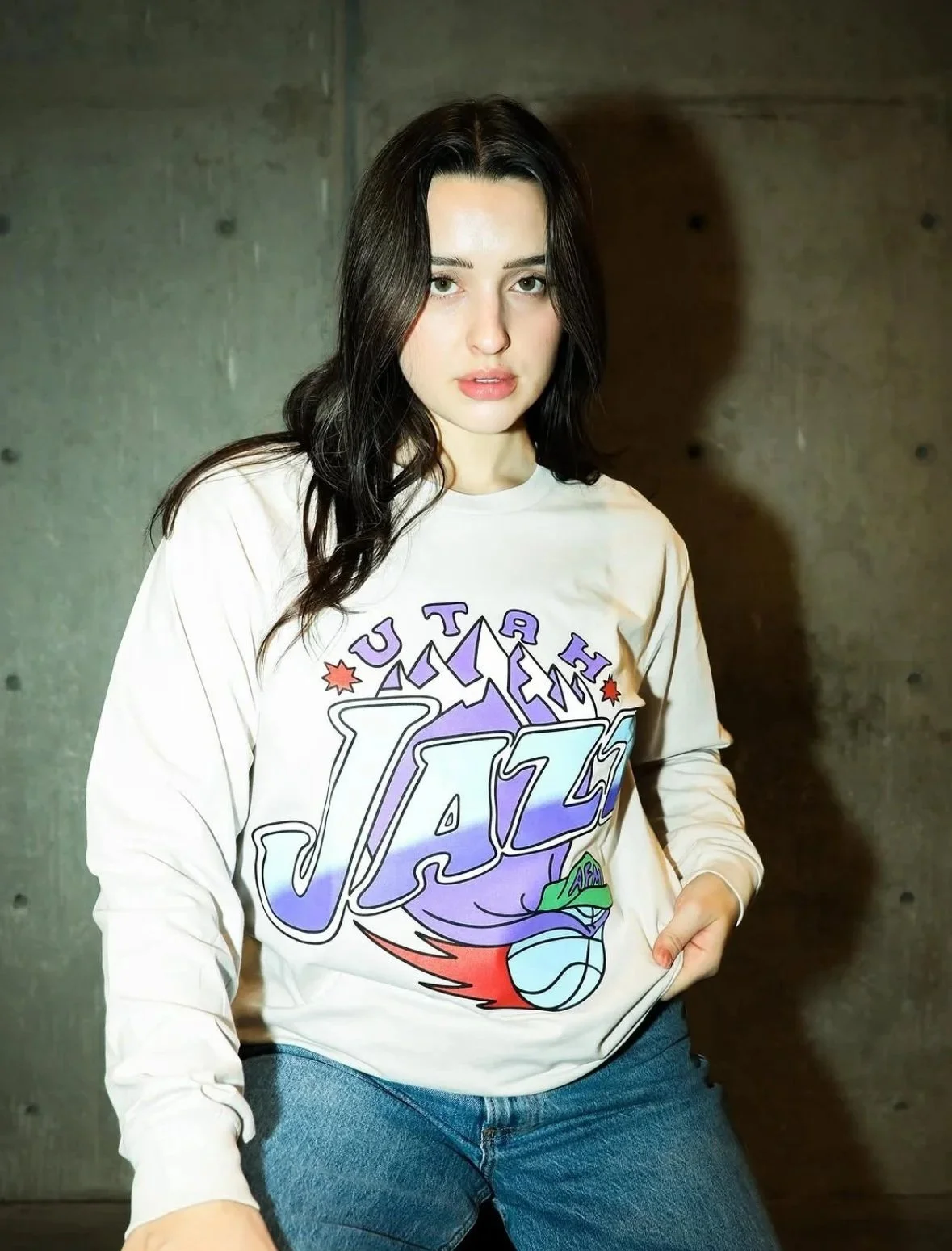 Young woman with dark hair wearing a cream long sleeve shirt with a colorful jazz-themed graphic design, and blue jeans, standing against a plain concrete wall.