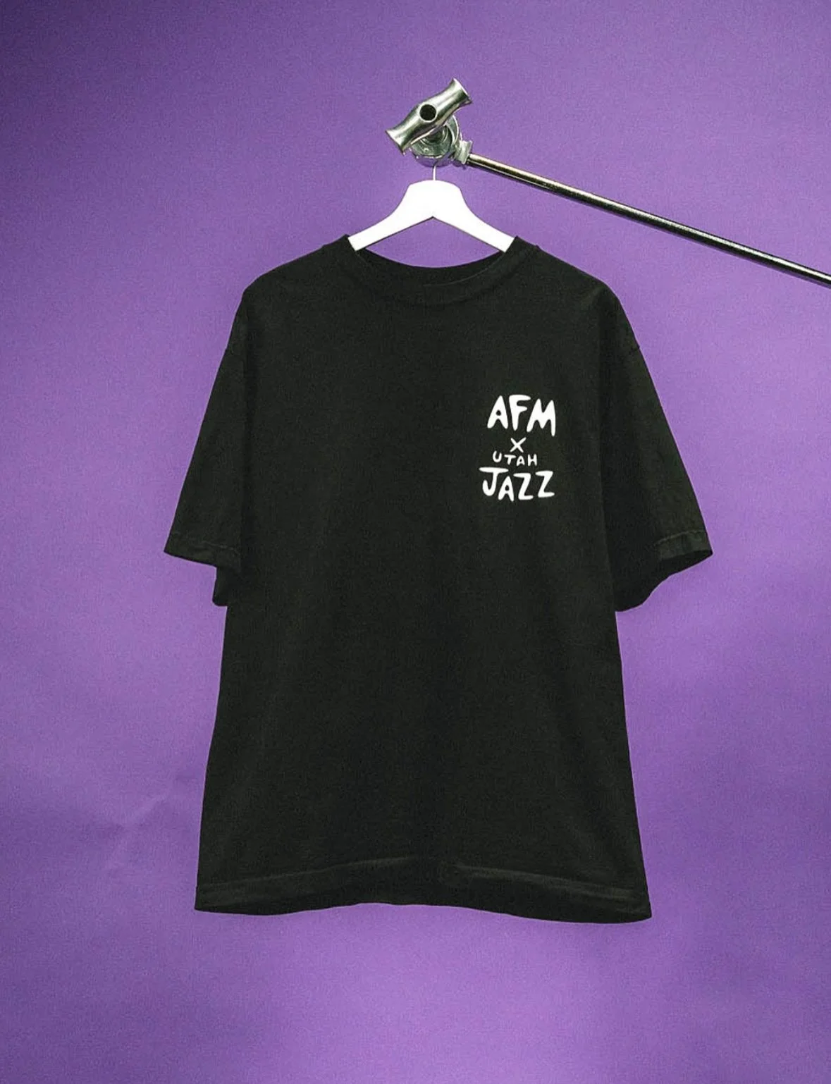 A black T-shirt with white text hanging on a white hanger against a purple wall. The text reads 'AFM x UTAH JAZZ' with a star symbol between 'AFM' and 'UTAH JAZZ'.