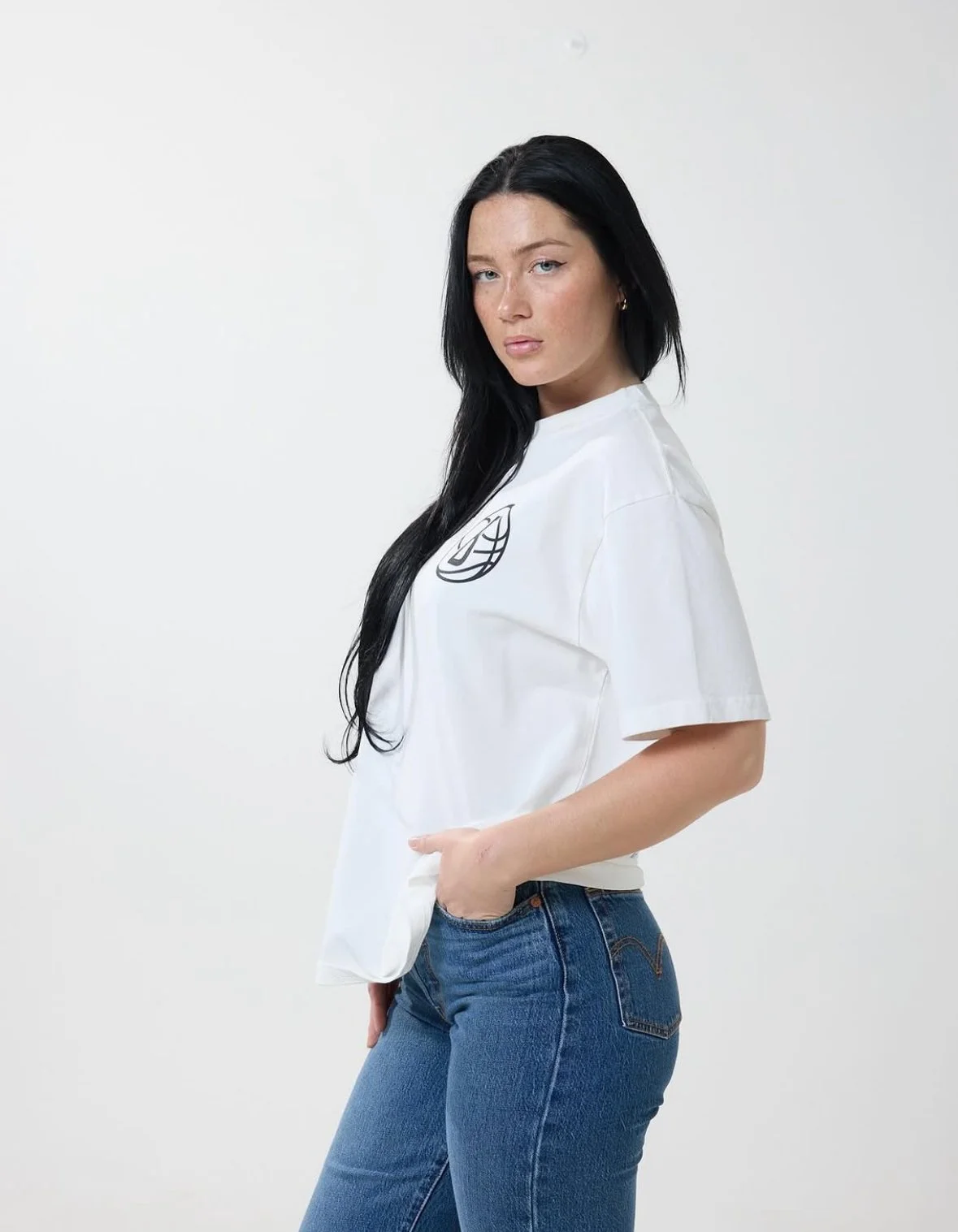 Young woman with long black hair wearing a white T-shirt with a graphic logo and blue jeans, standing against a plain white background.