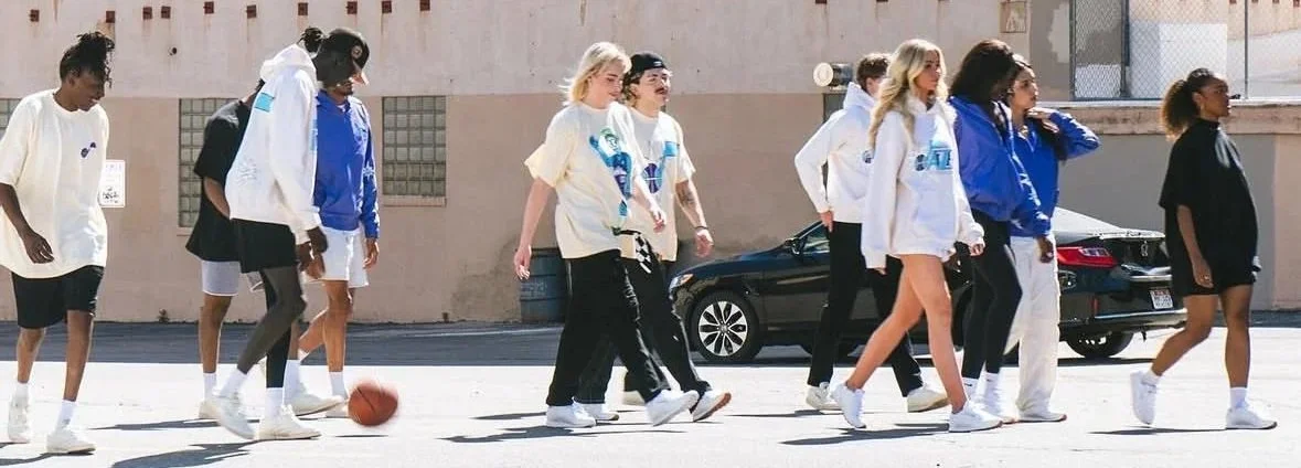 Group of young women walking on the street, some wearing casual outfits with hoodies and shorts, and one with a basketball.