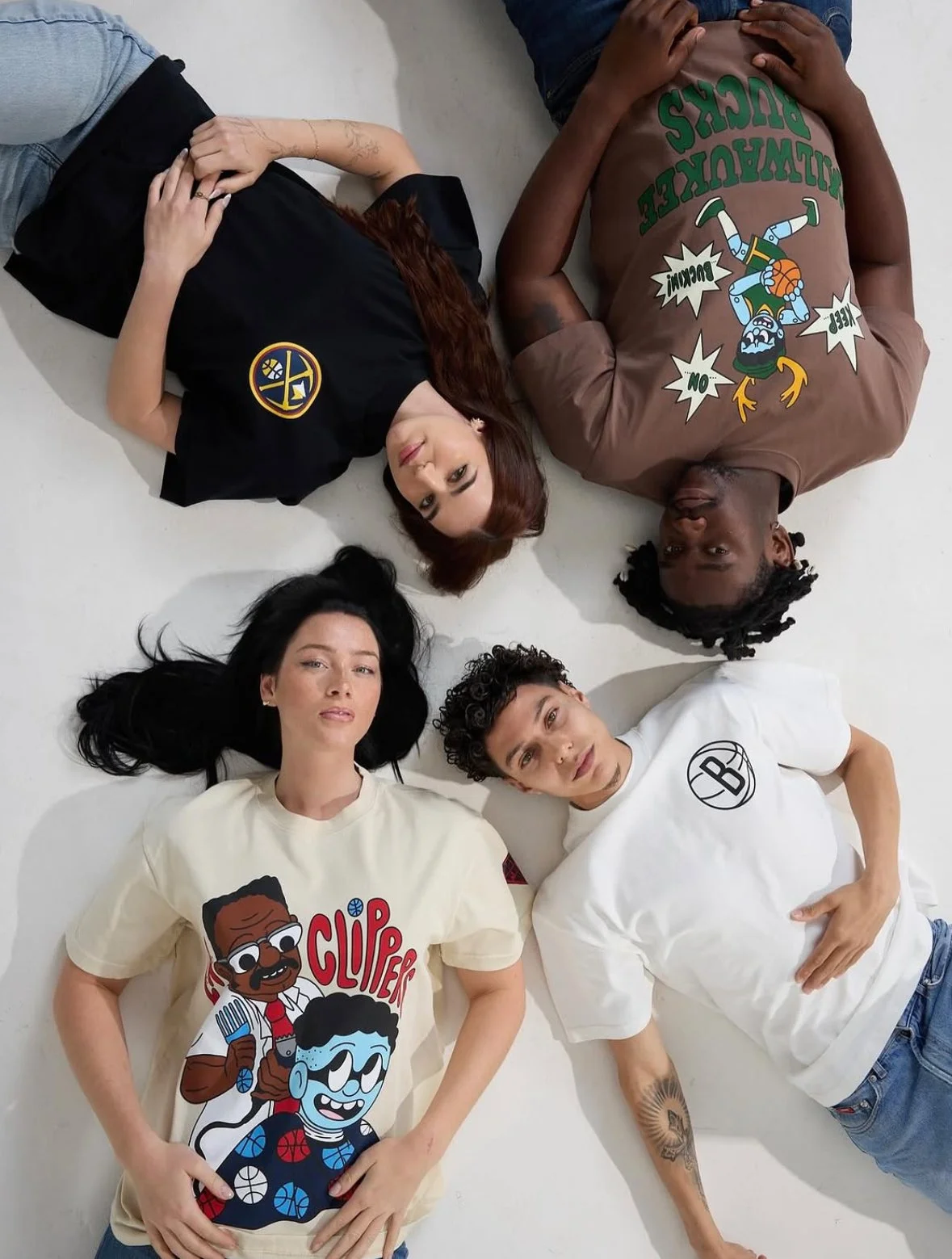 Group of five young people lying on white floor with heads close together, looking up at camera, each wearing graphic T-shirts.