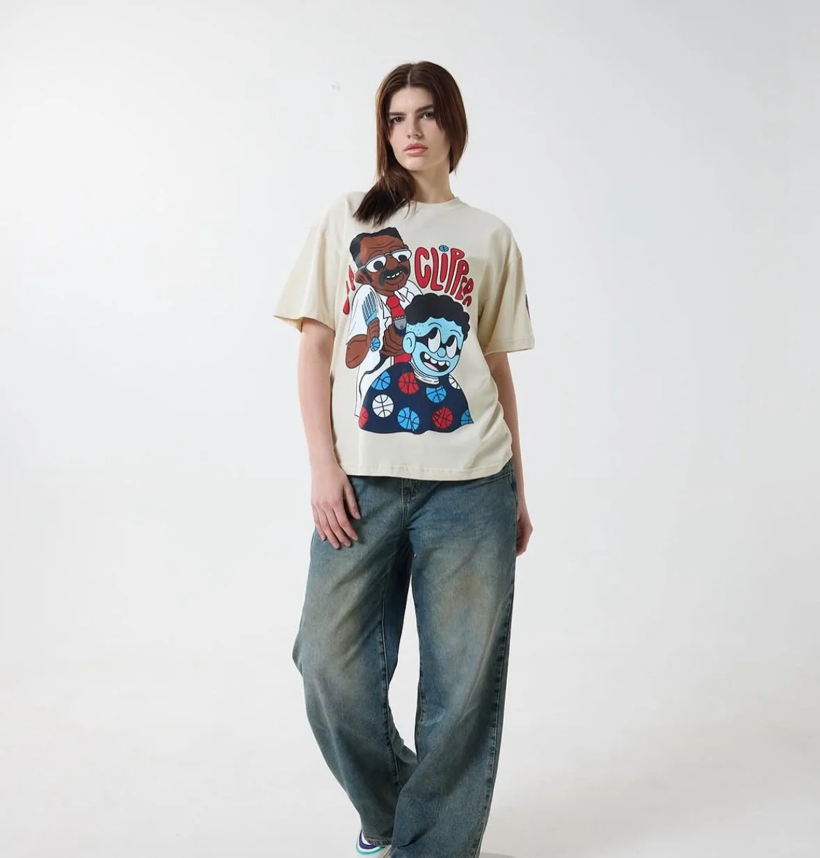 Young woman wearing a beige graphic t-shirt with cartoon characters and jeans, standing against a plain white background.