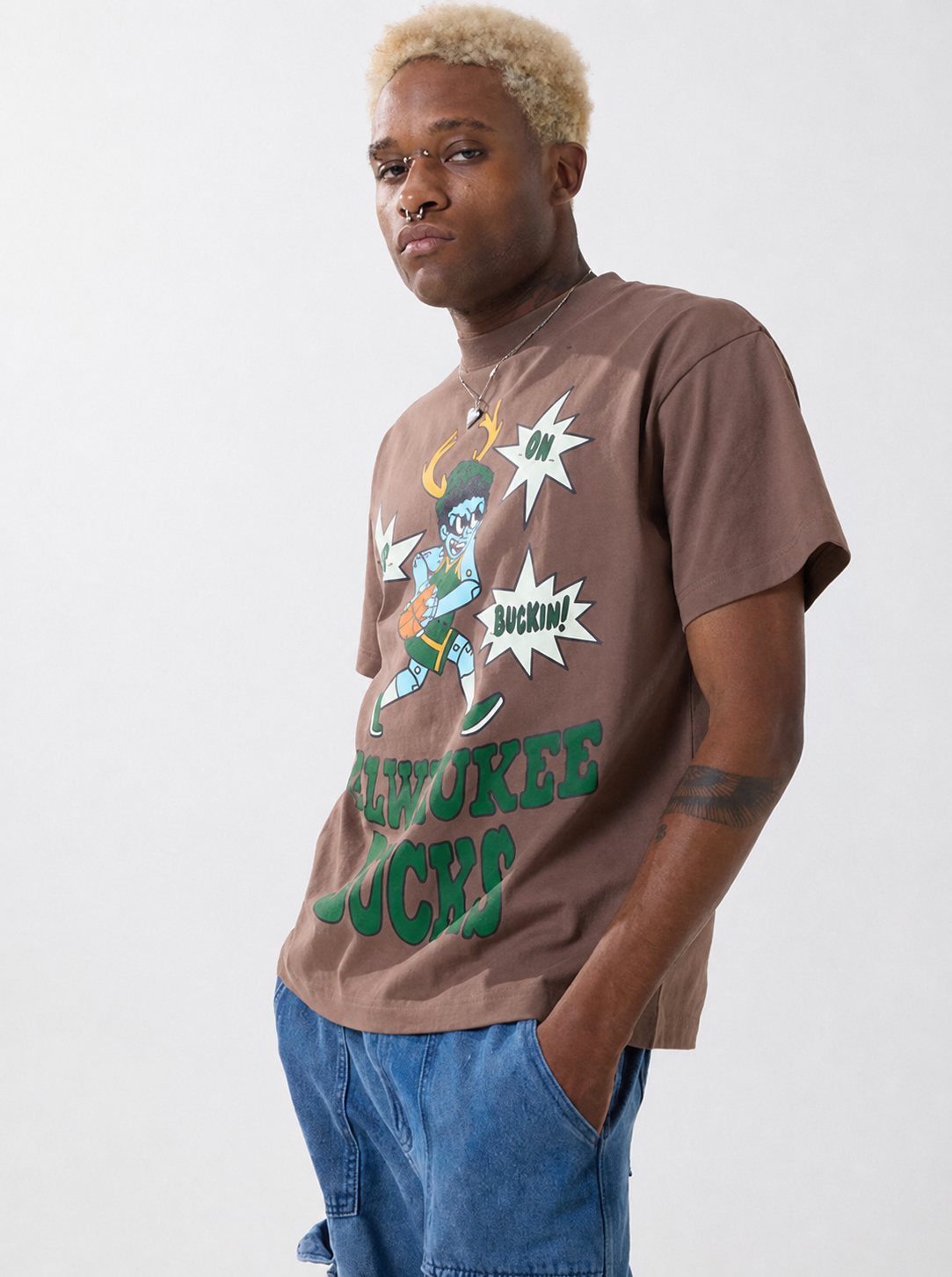 Young man with blond curly hair, tattoos, and nose piercings wearing a graphic T-shirt and blue jeans standing against a plain white background.