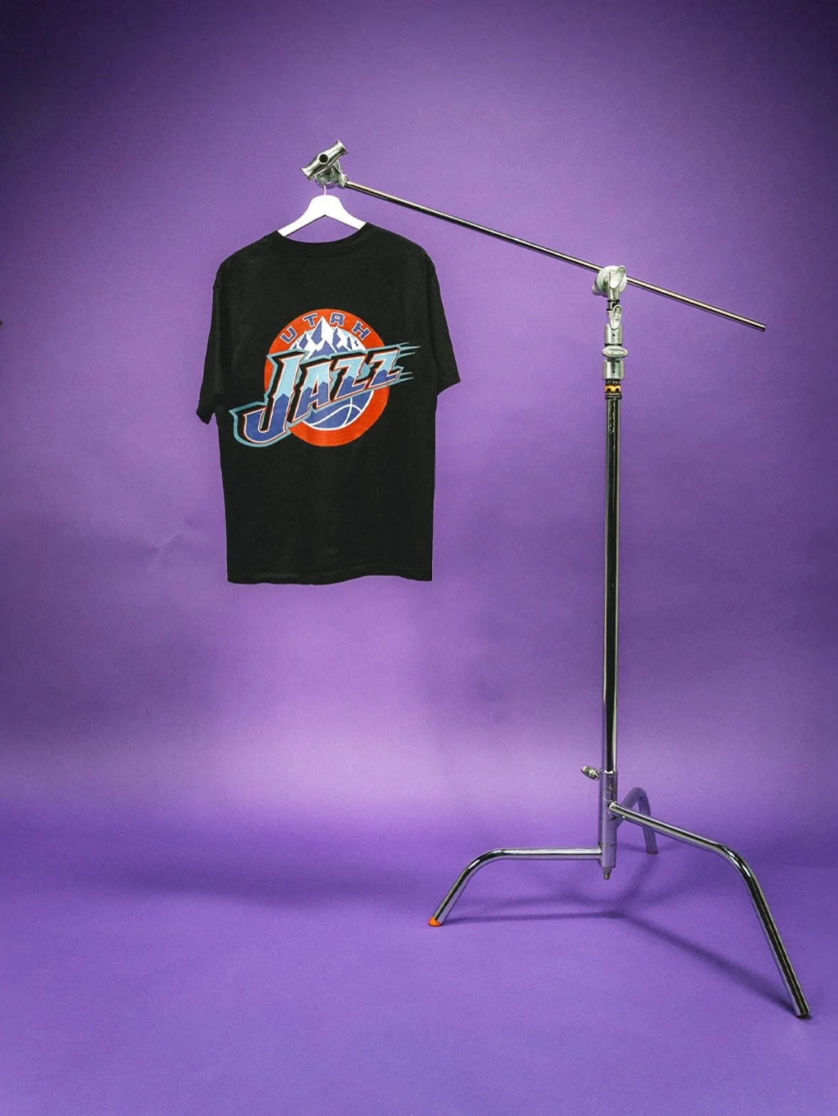 A black Utah Jazz t-shirt hanging on a white hanger, suspended on a metal arm stand against a purple background.