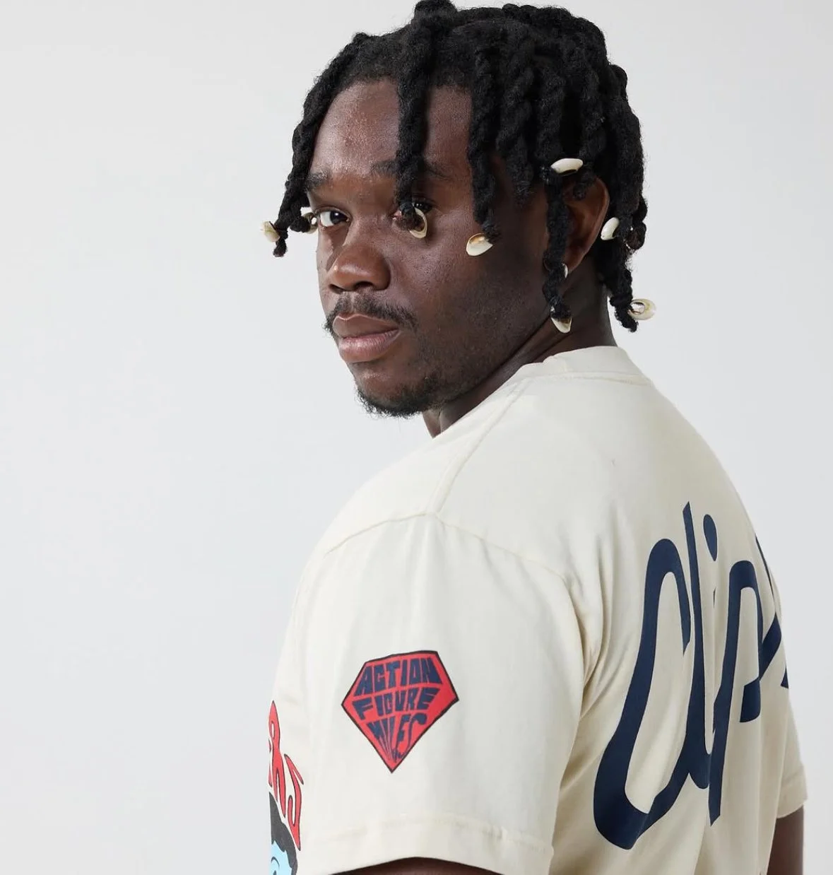 A man with styled dreadlocks attached with small white shells, wearing a beige t-shirt with patches, including one that says 'Action Figure' and a large signature on the back, against a plain white background.