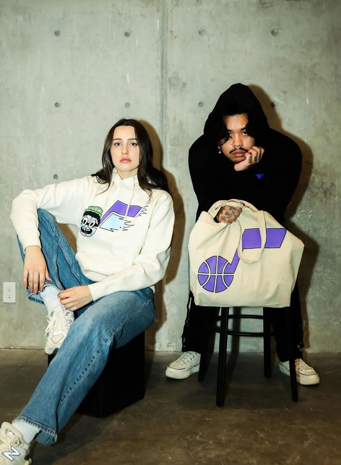 A woman sitting with one leg crossed in front of her and a man leaning on a chair with a bag, both in front of a concrete wall.