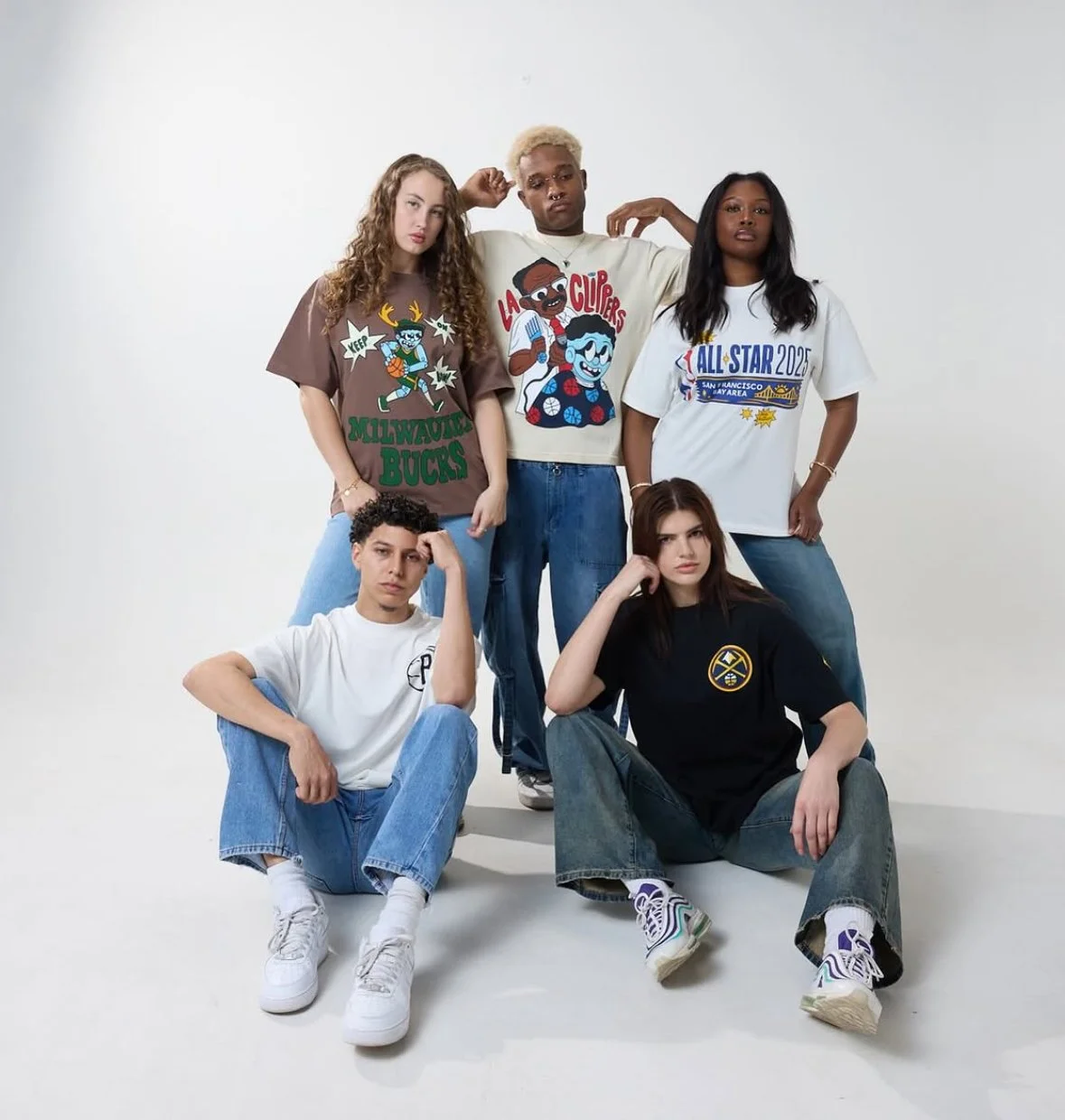 A diverse group of five young people posing together against a plain white background, wearing casual streetwear with graphic print T-shirts and sneakers.