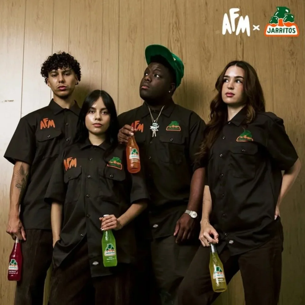 Four people, dressed in black work shirts with the AFM and San Antonio logo, stand in front of a wooden wall, holding jars of salsa and bottles of hot sauce, with the AFM x Jarritos logo in the top right corner.