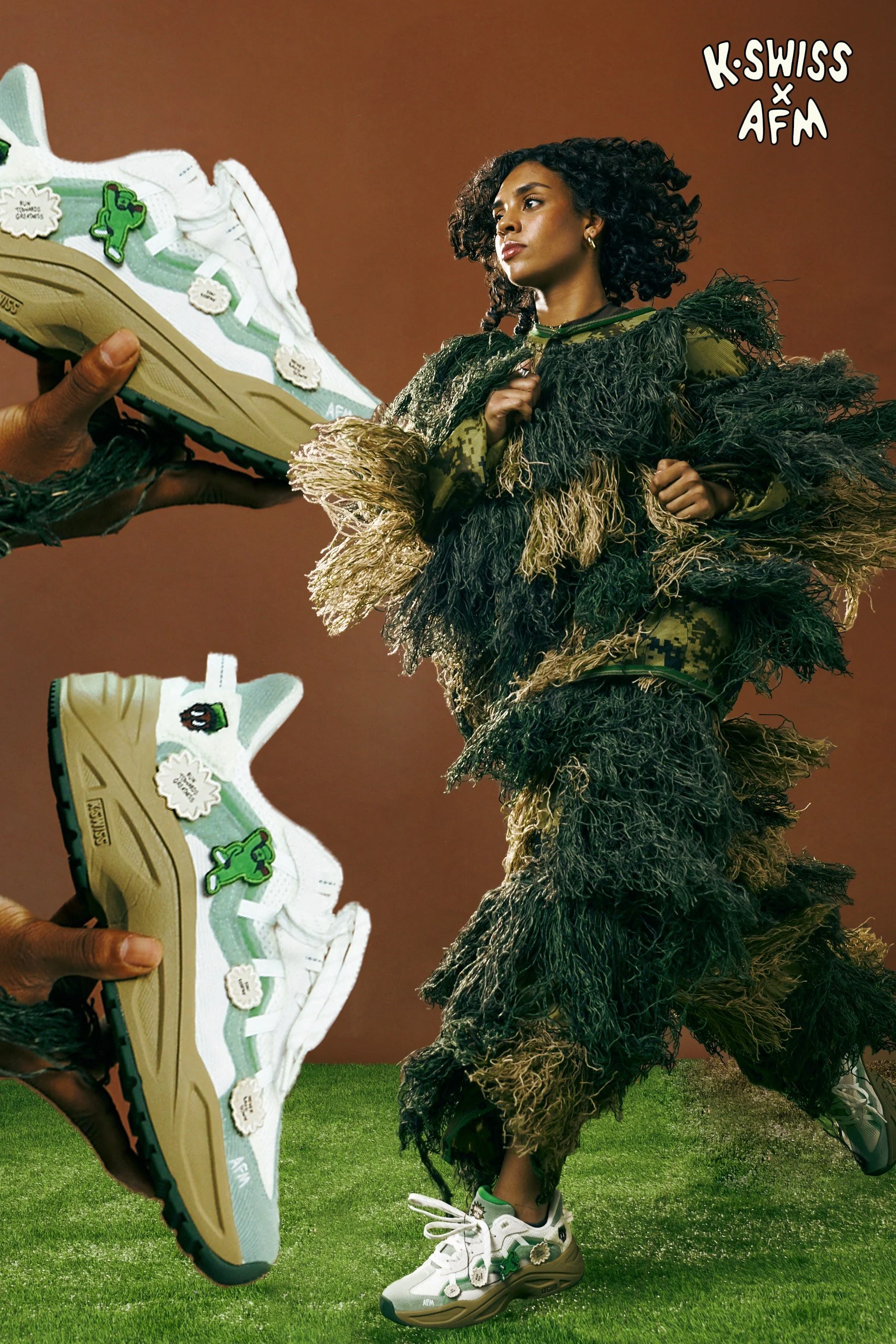 Model wearing a textured camouflage outfit with wide-legged pants and a fringed jacket, standing on green grass, holding two sneakers with white and green details decorated with cartoonish patches, in front of a brown background.