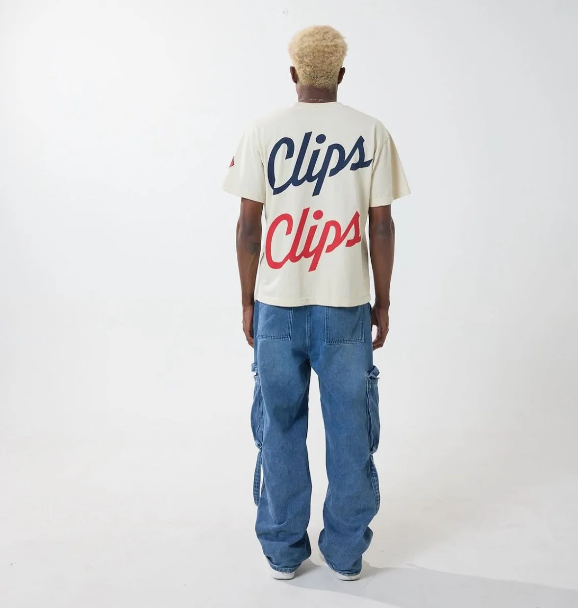 A person with bleached blonde hair, wearing a beige t-shirt with the word "Clips" written on the back in blue and red, blue jeans, standing against a plain white background, facing away from the camera.