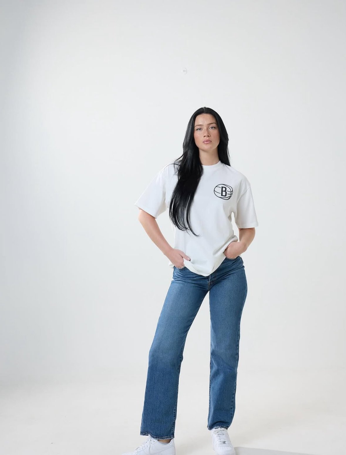 A woman with long black hair wearing a white t-shirt with a Brooklyn Nets logo and blue jeans, standing against a plain white background.
