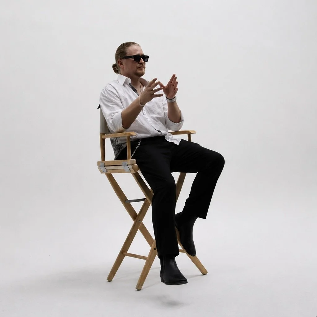 A man sitting on a wooden director's chair, wearing a white shirt, black pants, sunglasses, and black shoes, with a plain white background.