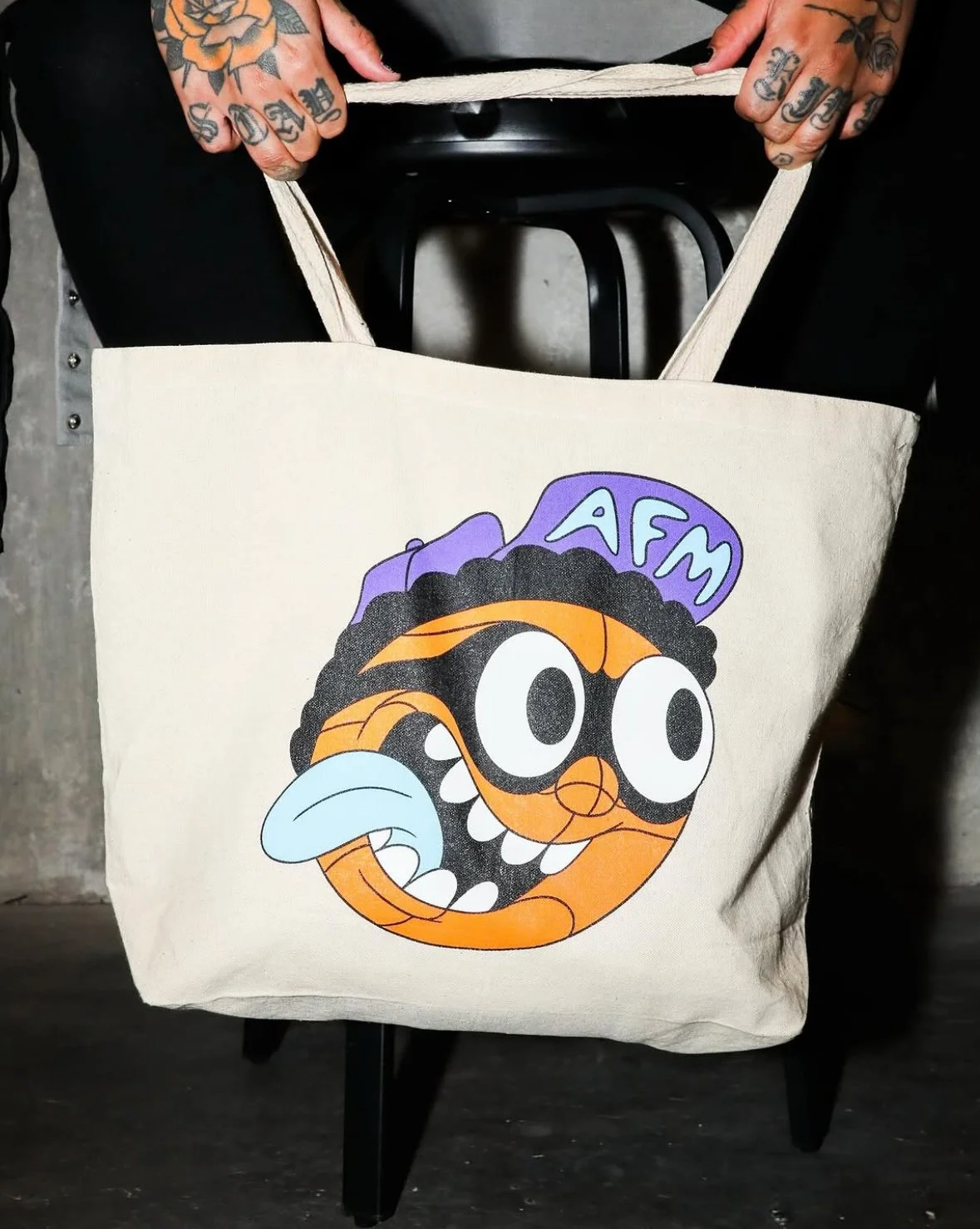 A beige tote bag with a colorful cartoon character printed on it, featuring large eyes, a wide open mouth with teeth, and a blue tongue. The character has black, curly hair and is wearing a purple hat with the letters 'AFM' on it. Someone with tattoos on their hands is holding the bag by its handles.