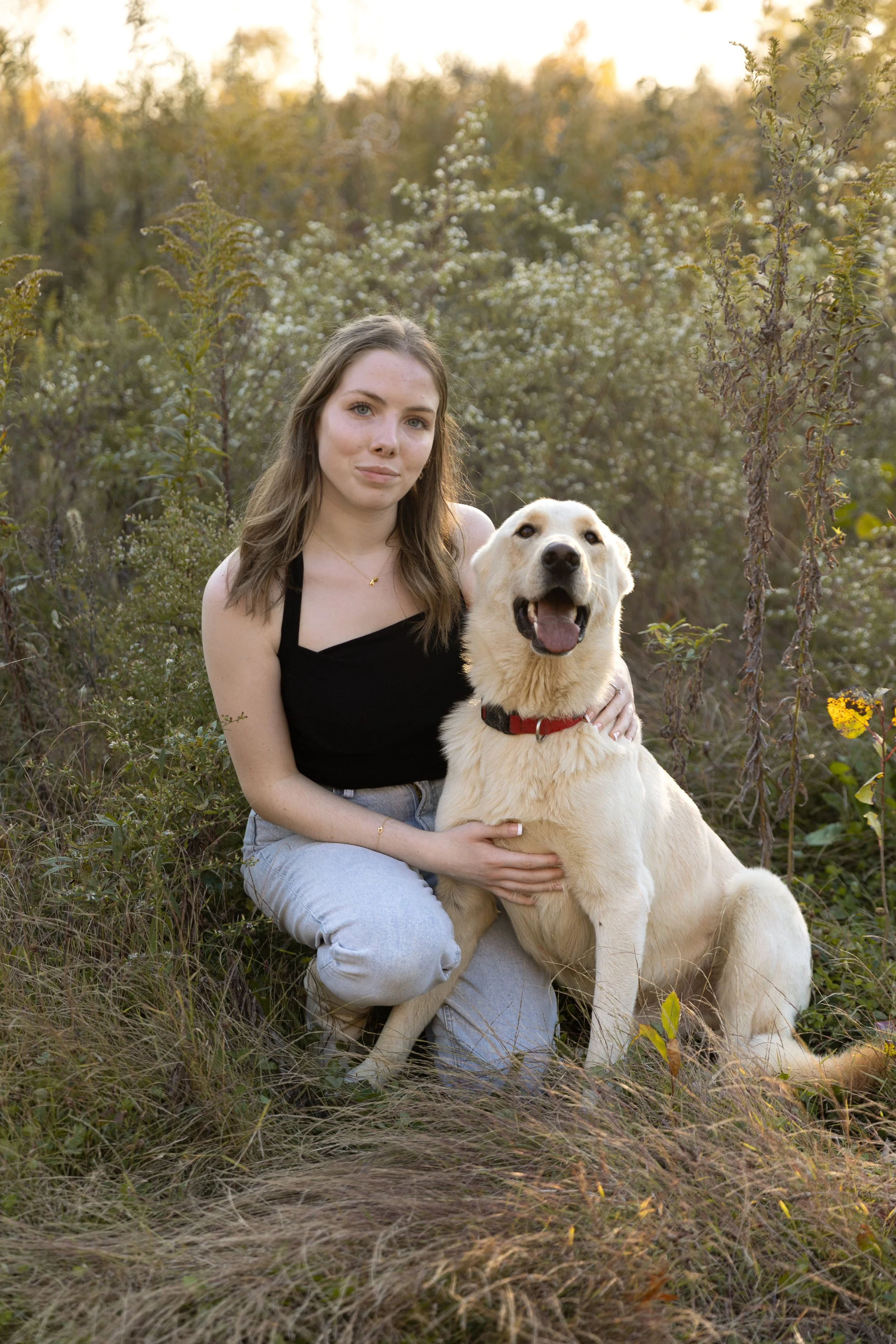 Young woman with brown hair wearing a black tank top and light blue jeans sitting outdoors on grass with a happy yellow Labrador retriever dog on a leash, surrounded by trees and shrubs in a natural setting during golden hour.