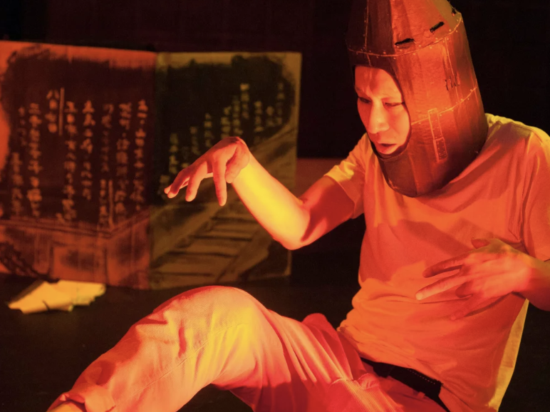 A portrait of a person with short black hair with blond highlights wearing a blue top, colorful earrings, and a necklace that says Queer AF