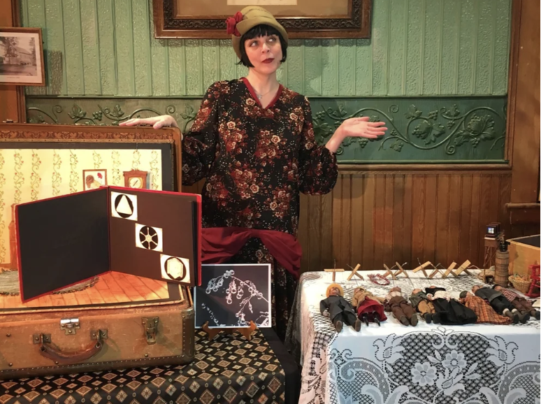 A selfie of a person with brown pigtail hair, clear glasses, smiling at the camera with a colorful puppet behind them
