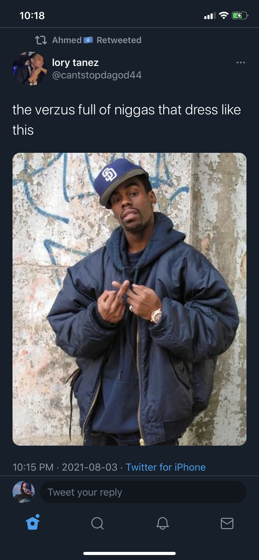 A young man wearing a baseball cap sideways, a dark jacket, and a wristwatch, standing against a graffiti-covered wall.