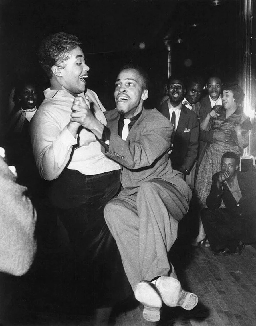 A joyful black couple dancing closely at a party, with other people smiling and watching them in the background.