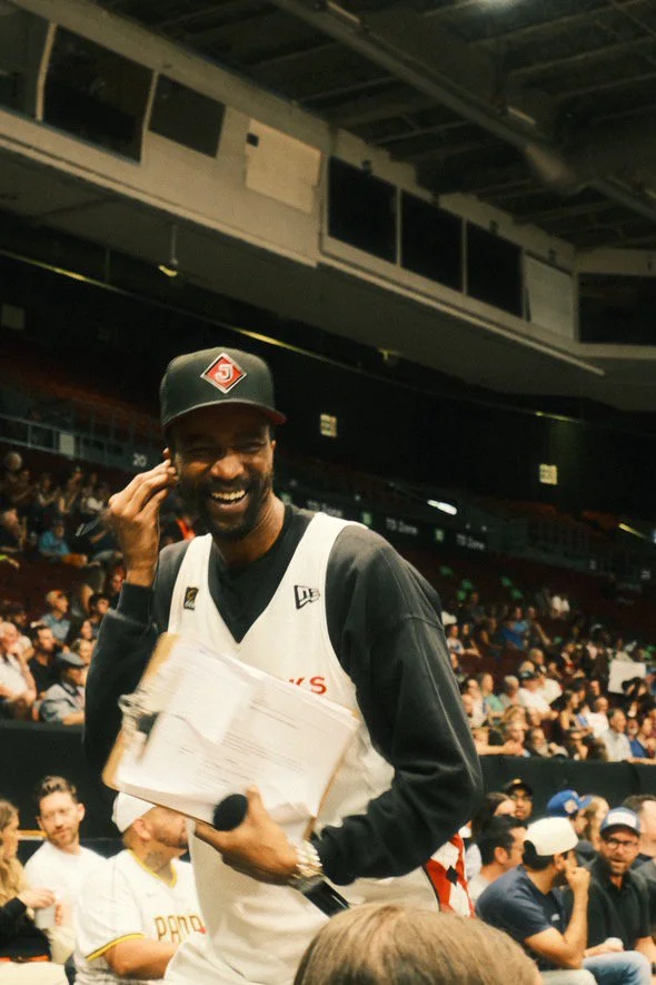 Bobby Brugal smiling and wearing a  Blackjacks sports jersey and cap, holding papers and a microphone at an indoor event with a crowd in the background.