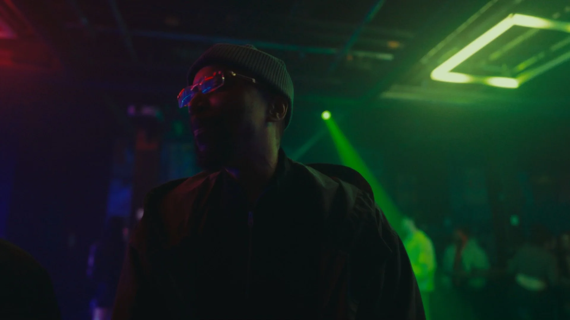A person wearing a grey beanie and glasses with colorful reflections, in a dimly lit club with green and yellow neon lights.