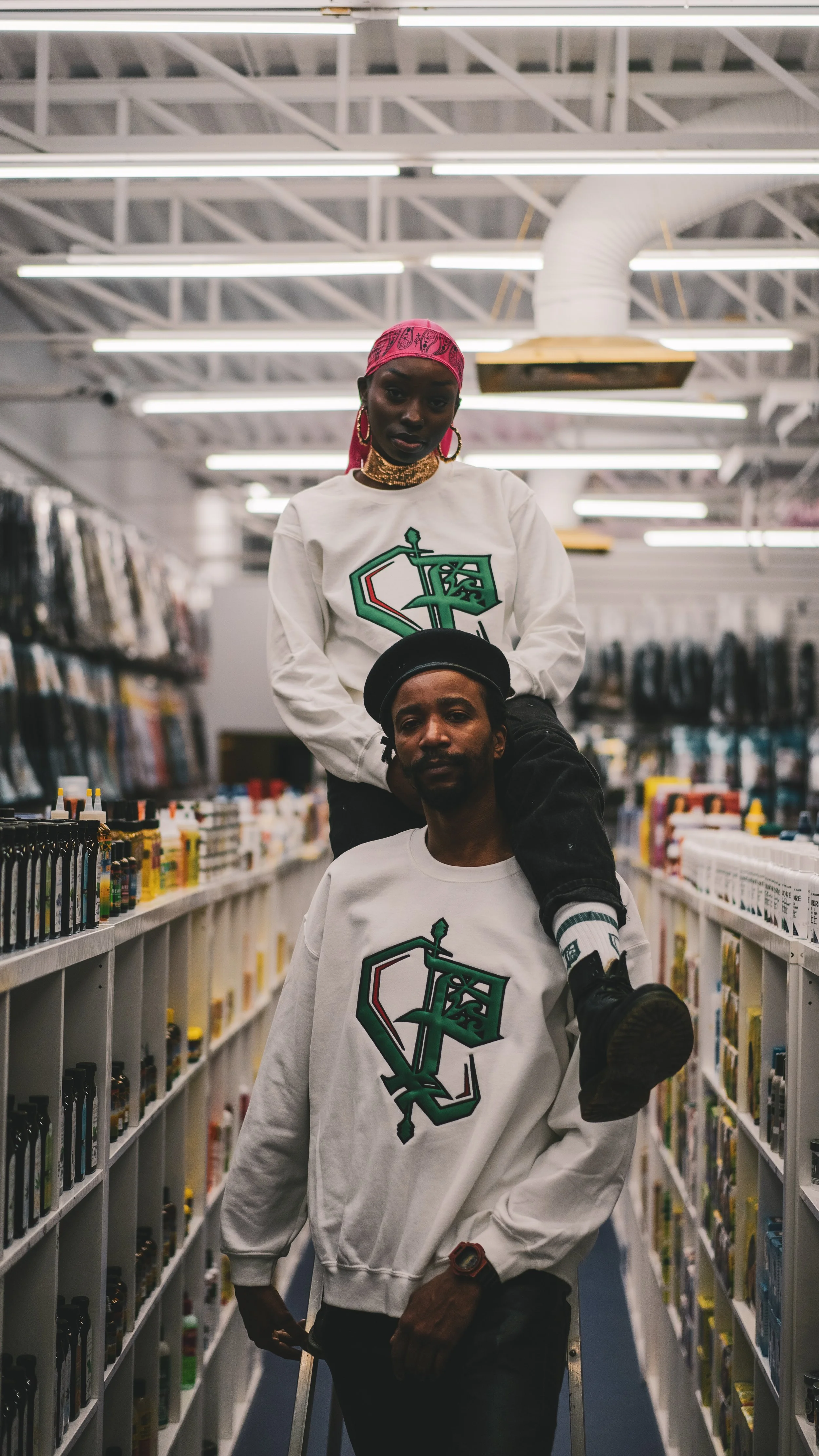 Two young Black individuals in a store aisle, both wearing matching white sweatshirts with a green ornate sword and shield logo. The person on the floor is carrying a shopping cart, and the person sitting on their shoulders is wearing a pink bandana 