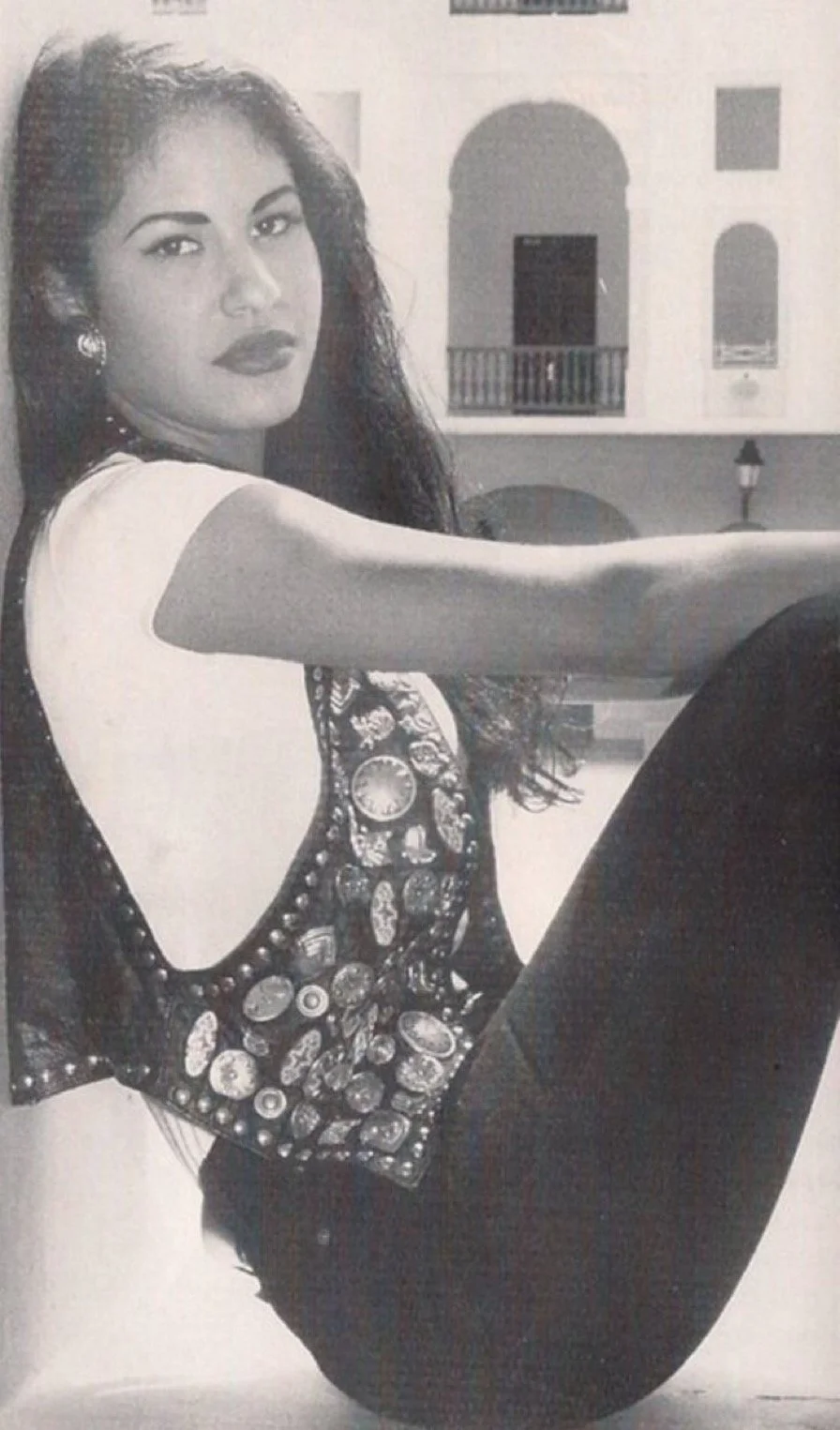 Black and white photo of a woman with dark wavy hair, dark lipstick, and earrings, sitting with her legs bent and her arm extended in front of a building with arched windows and balconies.
