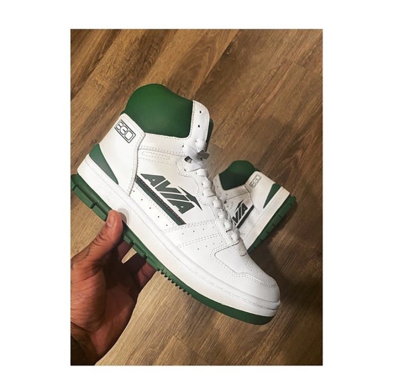 A pair of white and green high-top sneakers with the AVIA logo on the side, held by a person's hand against a wooden floor background.