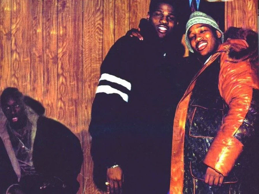 Two men standing close together, smiling, one with a black hoodie with white stripes, the other wearing a warm orange coat and a green knit hat. A third man is sitting nearby, dressed in dark clothing, against a wooden wall background.