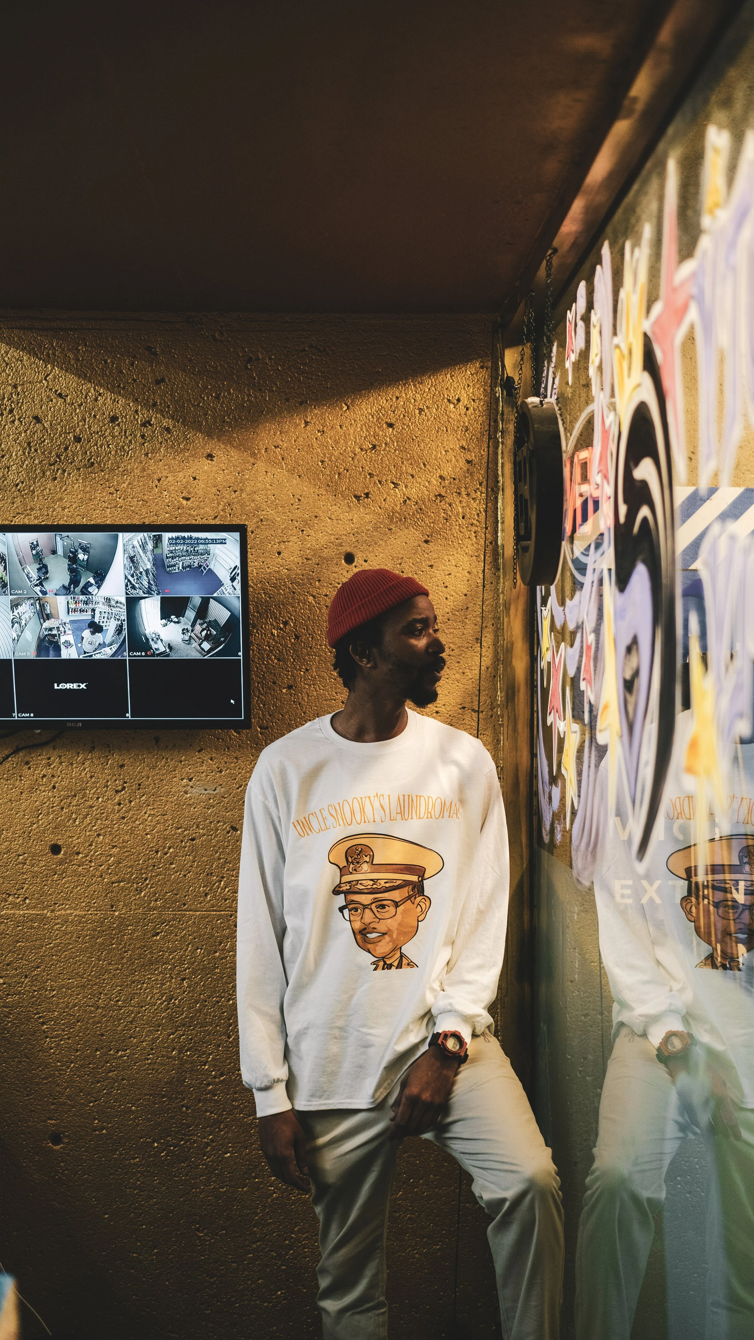A man with a red beanie and white sweatshirt standing in front of a wall with colorful artwork, with a mirror reflecting part of his outfit and a surveillance monitor showing multiple camera feeds on the wall.