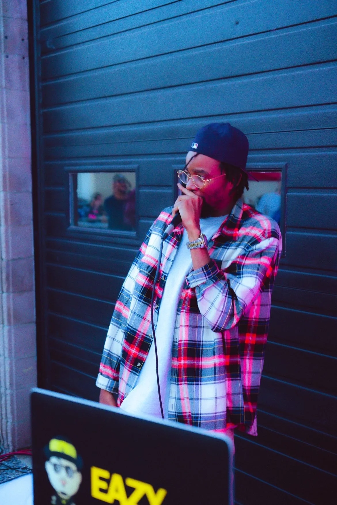 A young man wearing glasses, a black baseball cap backwards, a white t-shirt, and a red plaid shirt, standing against a black wall with small windows, holding a microphone near his mouth, with a laptop in front of him that has a sticker reading "EAZY