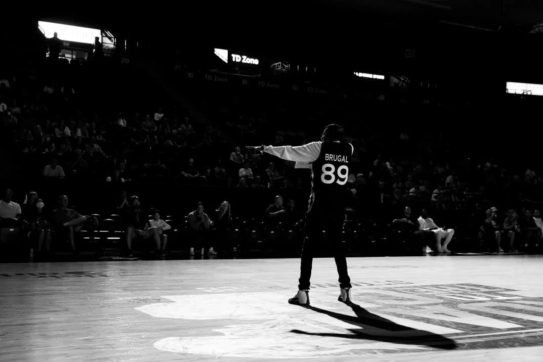 A basketball player with the jersey number 89 and the name 'Brugal' is standing on the court, pointing with his right arm, while the audience watches from the bleachers in a dimly lit arena.